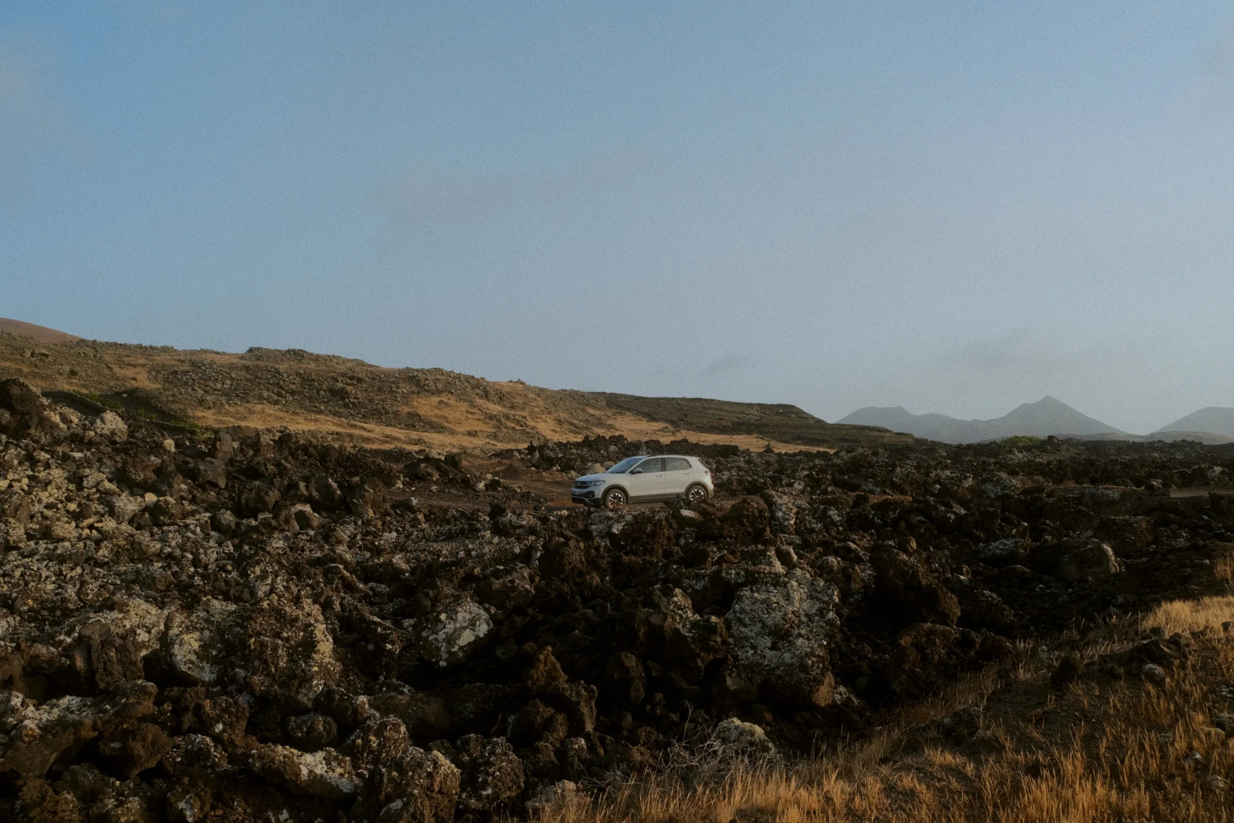 Une voiture blanche stationnée sur un terrain rocheux sous un ciel dégagé, avec des montagnes en arrière-plan.
