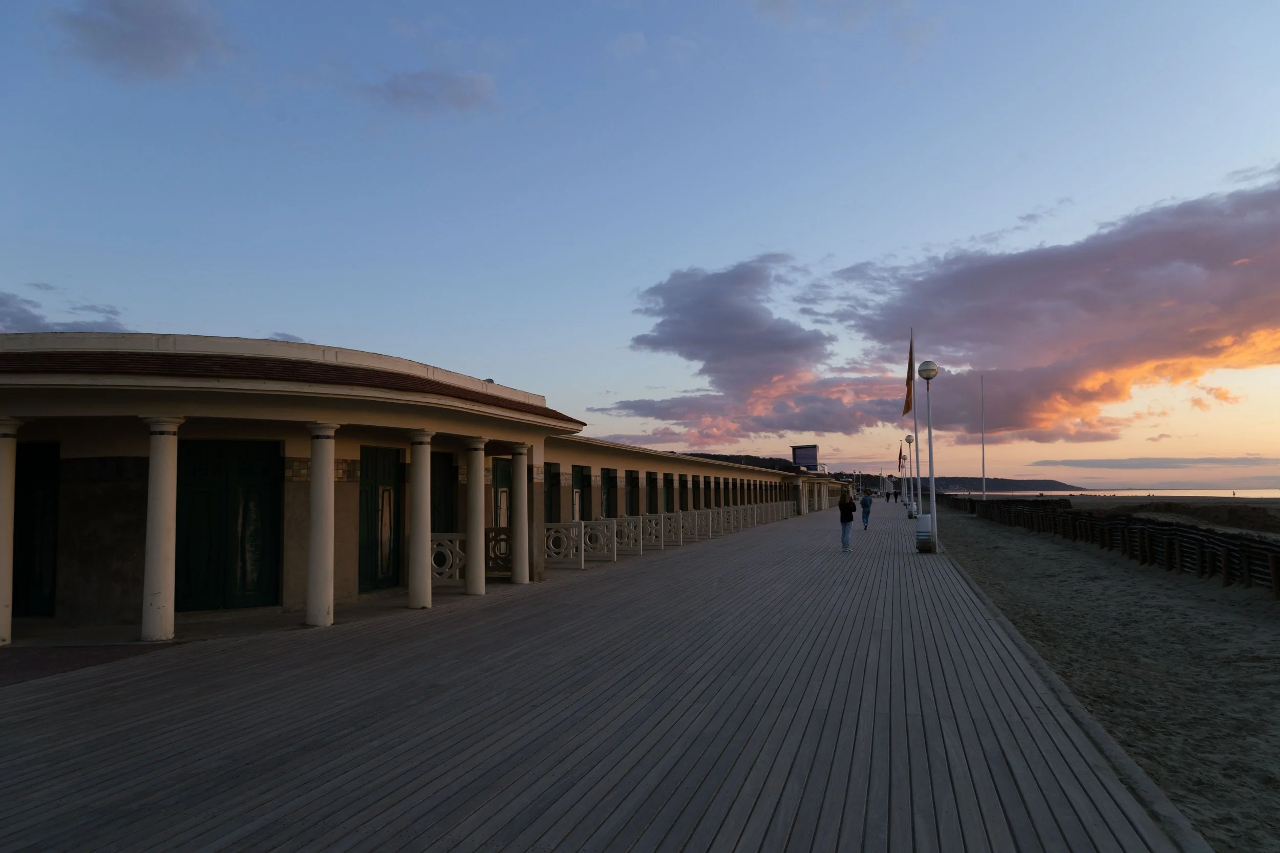 Promenade en bord de mer au coucher du soleil avec bâtiment à colonnes, lampadaires, drapeaux et ciel coloré.