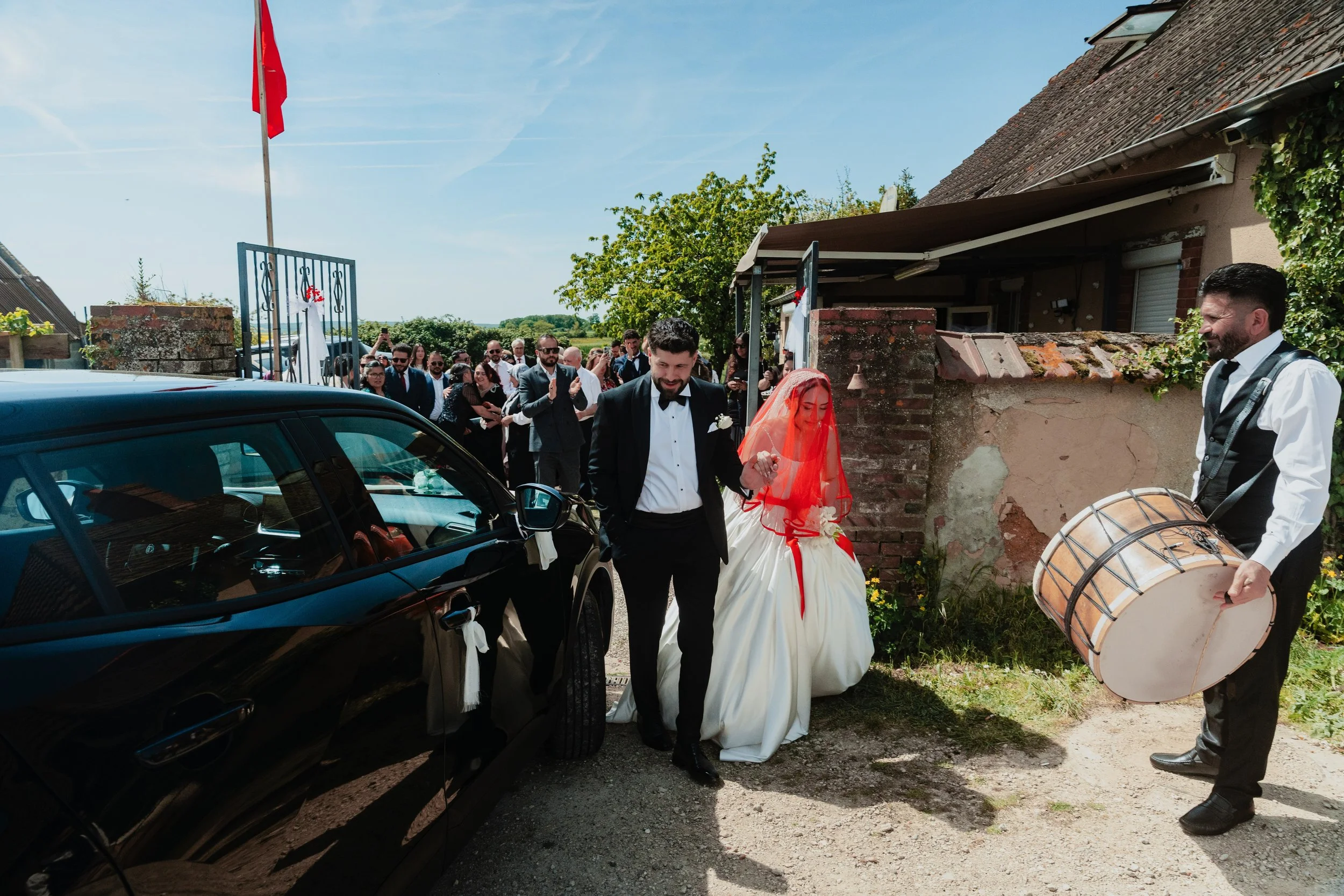 Un groupe de personnes en tenue de mariage, dont une femme en robe de mariée avec un voile rouge, se rassemblent devant une maison en pierre. Un homme en costume noir avec un nœud papillon semble accompagner la mariée vers une voiture noire, tandis q