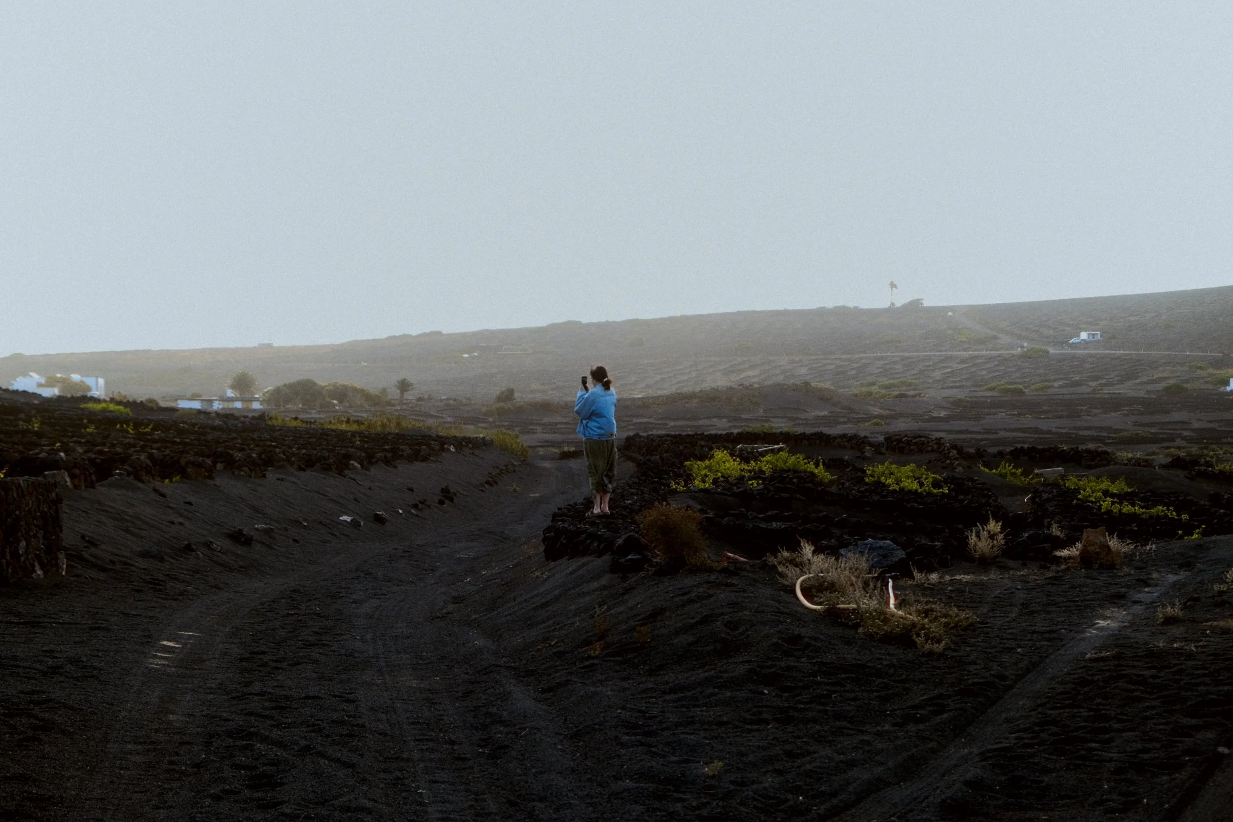 Une personne en veste bleue prend une photo avec un smartphone dans un paysage volcanique sombre avec un ciel gris nuageux, des plantations en toile de fond et une route visible.