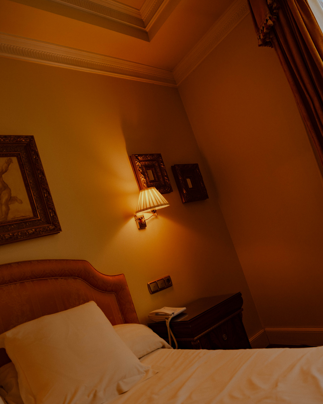 Chambre d'hôtel avec un lit, une lampe de chevet allumée, un tableau encadré, une vieille commode avec un téléphone et des prises électriques, rideaux en tissu.