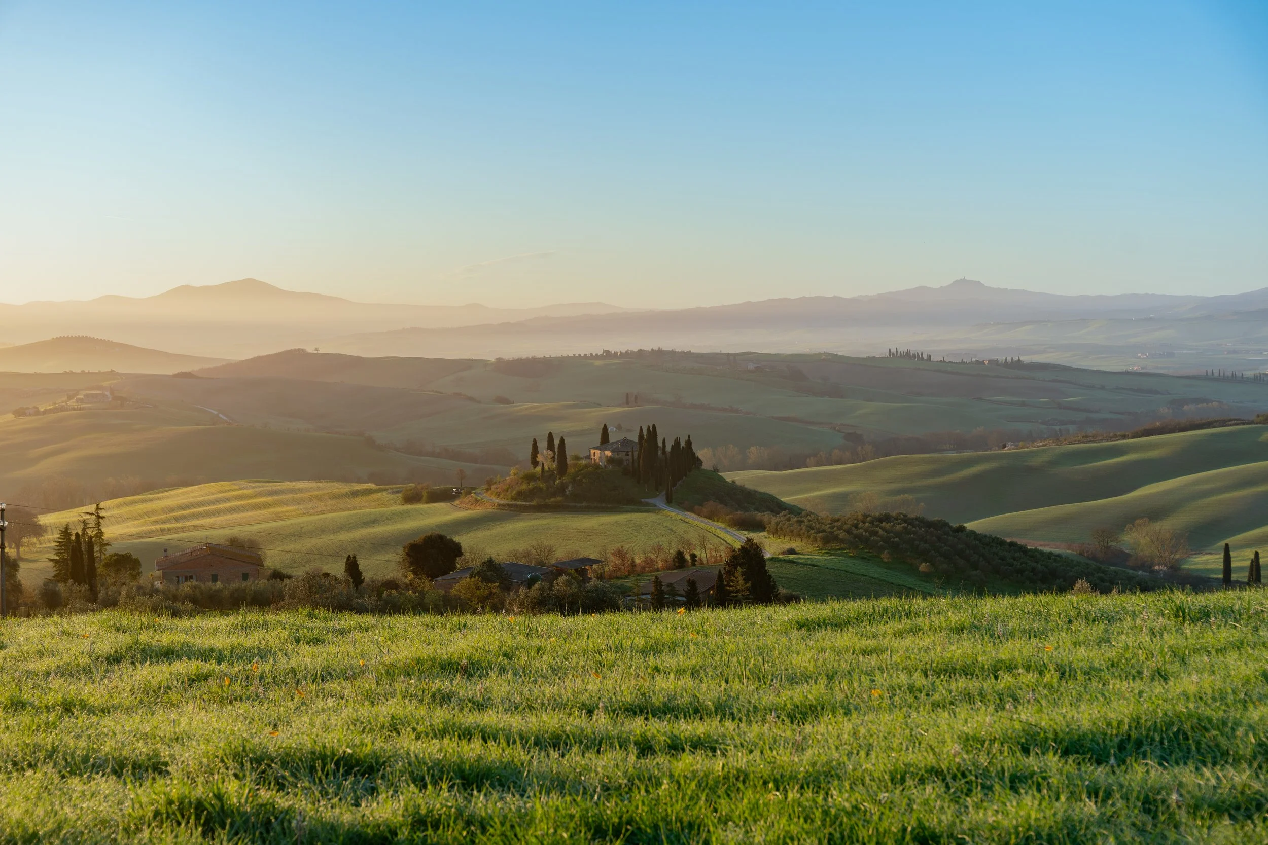 Paysage de collines verdoyantes sous un ciel clair avec quelques montagnes à l'arrière-plan et un petit groupe de maisons entourées d'arbres.