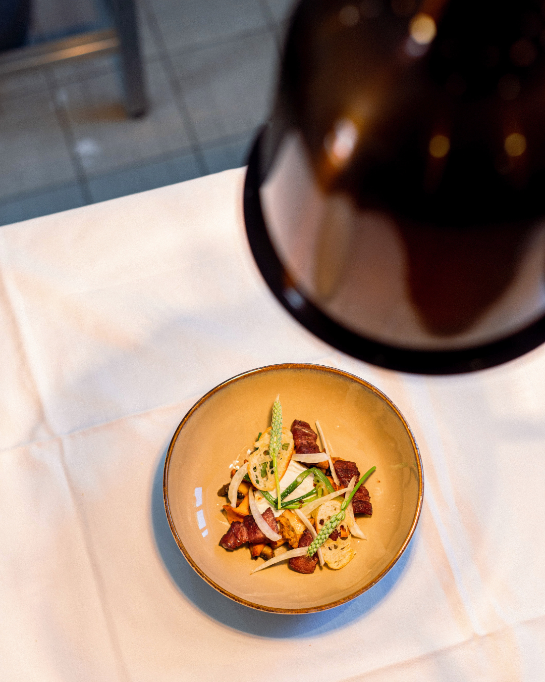 Une assiette de garniture, avec des morceaux de viande, des légumes et des herbes, vue d'en haut sur une table blanche, avec un pot en verre sombre au-dessus.
