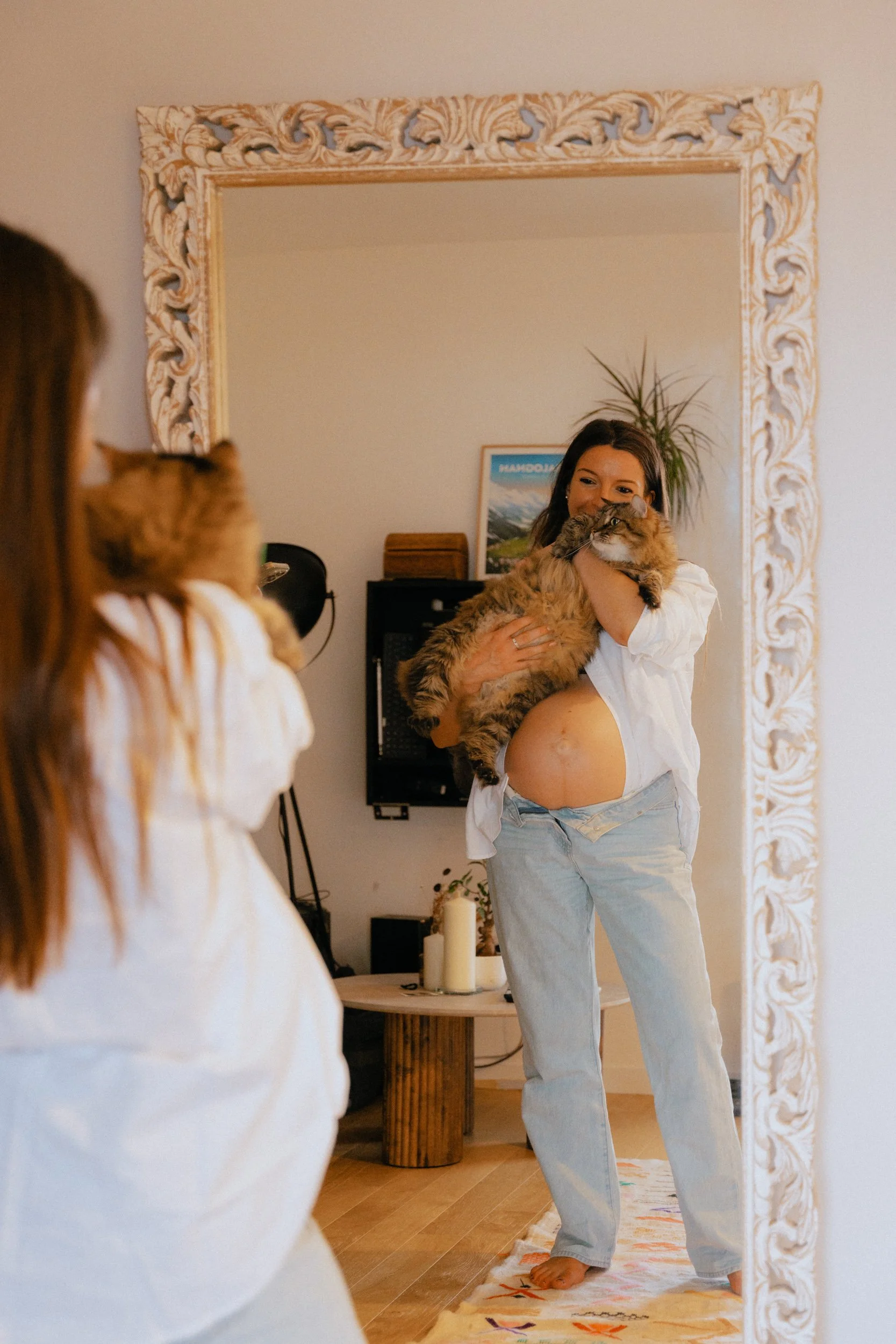 Une femme souriante tient un chat tigré et blanc devant un miroir, dans un intérieur lumineux avec des décorations naturelles et une peinture de paysage en arrière-plan, dans une pièce chaleureuse.