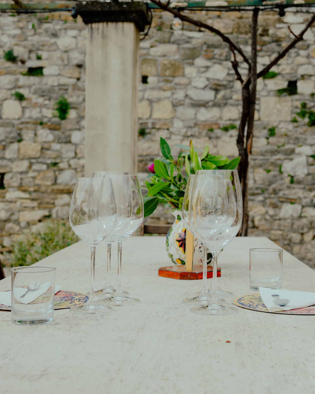 Table dressée avec des verres à vin, un vase avec des fleurs vertes et roses, et des assiettes avec des cuillères, en extérieur contre un mur en pierre.