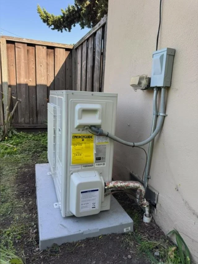 Outdoor heat pump compressor unit on a concrete slab next to a house wall with electrical panels and conduit.