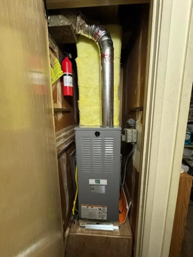 A furnace installed in a small closet with yellow insulation behind it, a fire extinguisher on a shelf, and a vent pipe connecting to the ductwork.