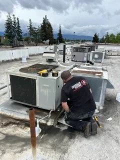 A technician working on air conditioning units on a building rooftop, with trees and a cloudy sky in the background.