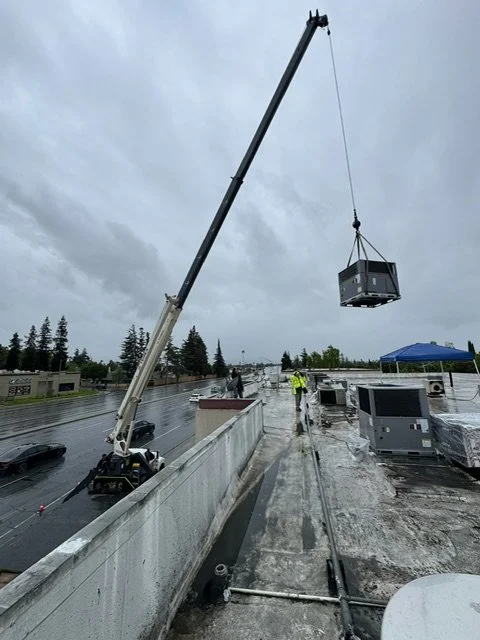 A crane lifting a large HVAC unit onto a rooftop parking lot during overcast weather, with workers in safety vests and a tent in the background.