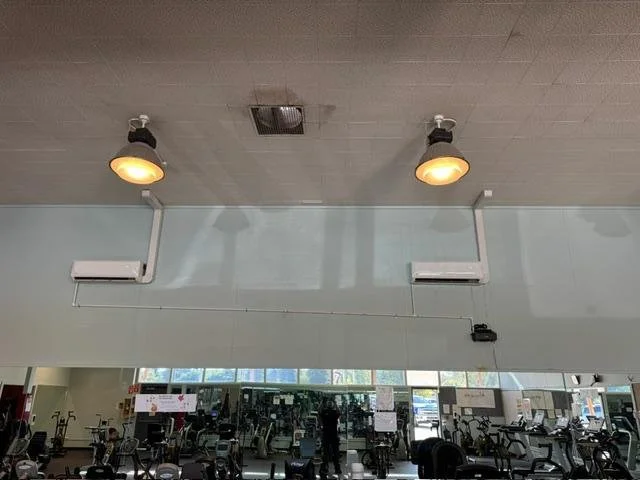 View of a gym's ceiling with two ceiling lights, air conditioning units, and exercise equipment in the background.