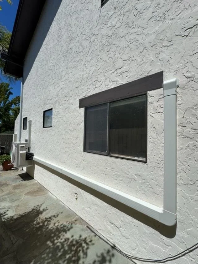 Exterior wall of a house with white stucco finish, featuring a sliding window with a brown frame, a white gutter pipe along the wall, and an air conditioning unit outside.