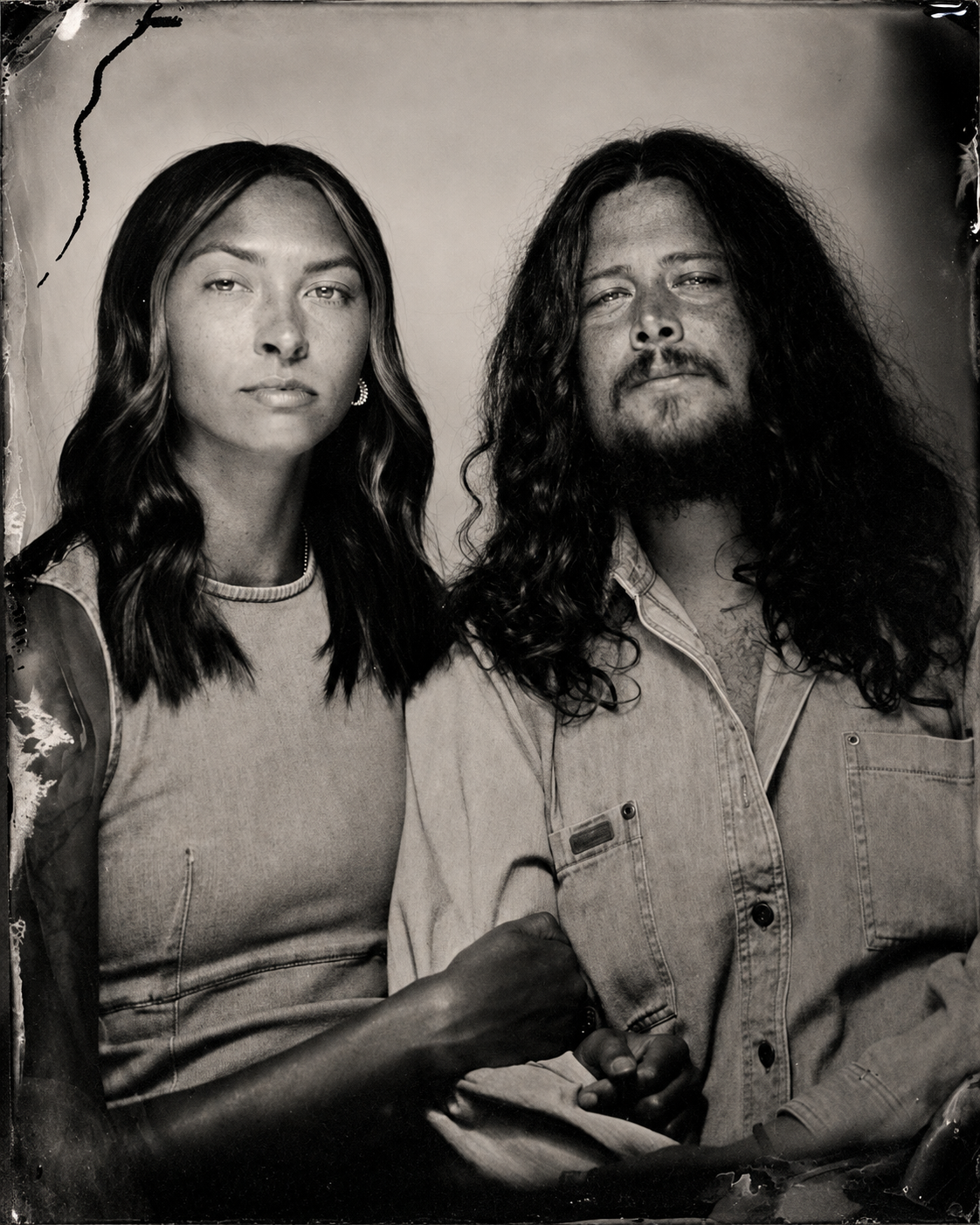 Black and white portrait of a young woman with long wavy hair and a young man with long curly hair and a beard, both wearing casual clothing, looking at the camera.