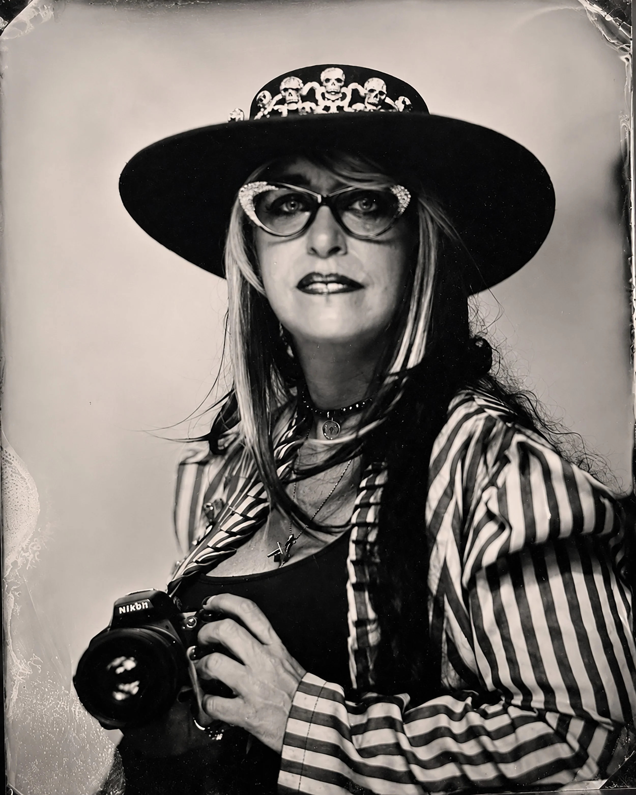 A woman taking a selfie with a DSLR camera, wearing a wide-brimmed hat decorated with skulls, glasses with rhinestones, a striped shirt, and layered necklaces, against a plain background.  TINTYPE PORTRAIT