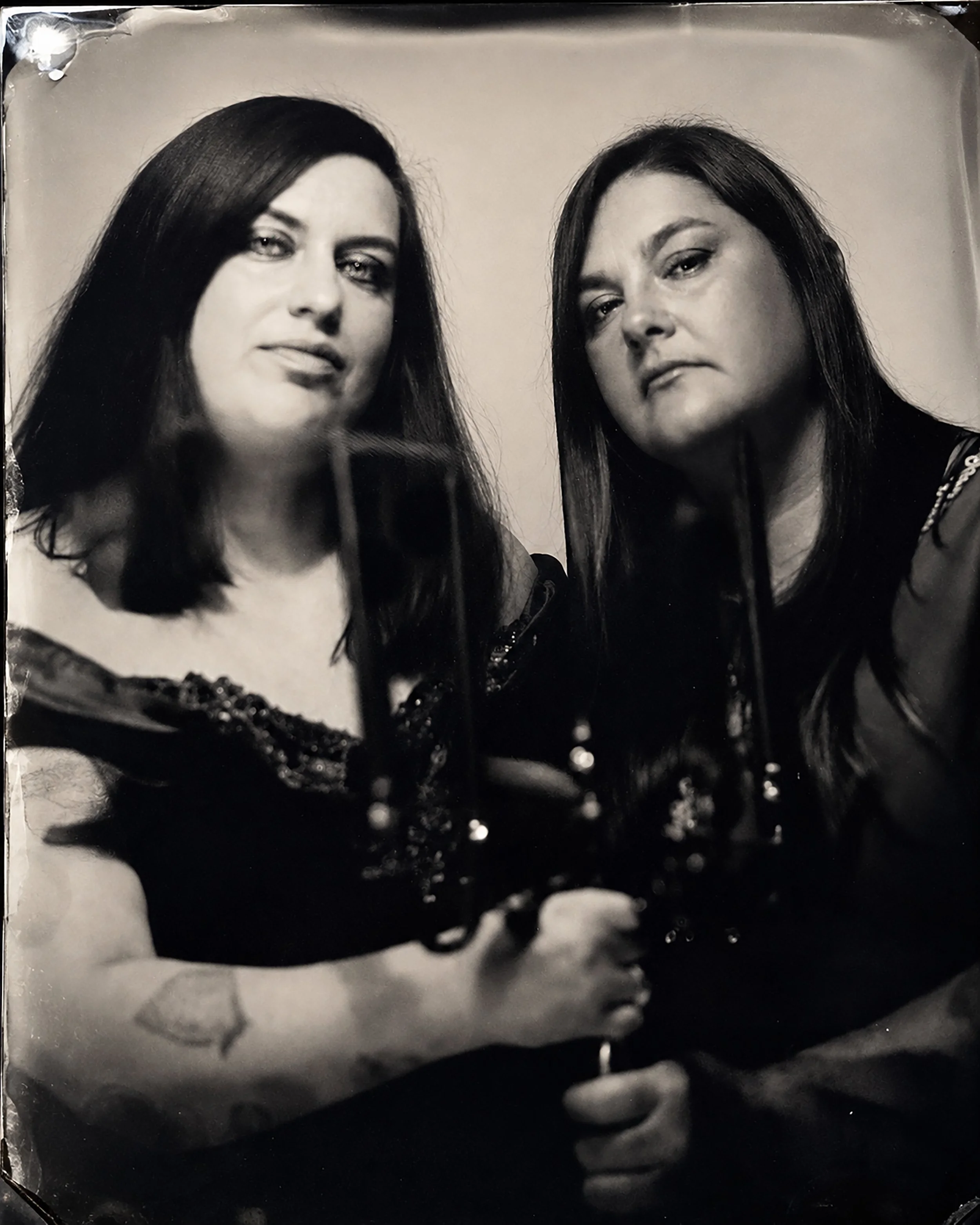 A black and white portrait photograph of two women with dark hair, looking at the camera with serious expressions, one on the left and one on the right. TINTYPE PORTRAIT