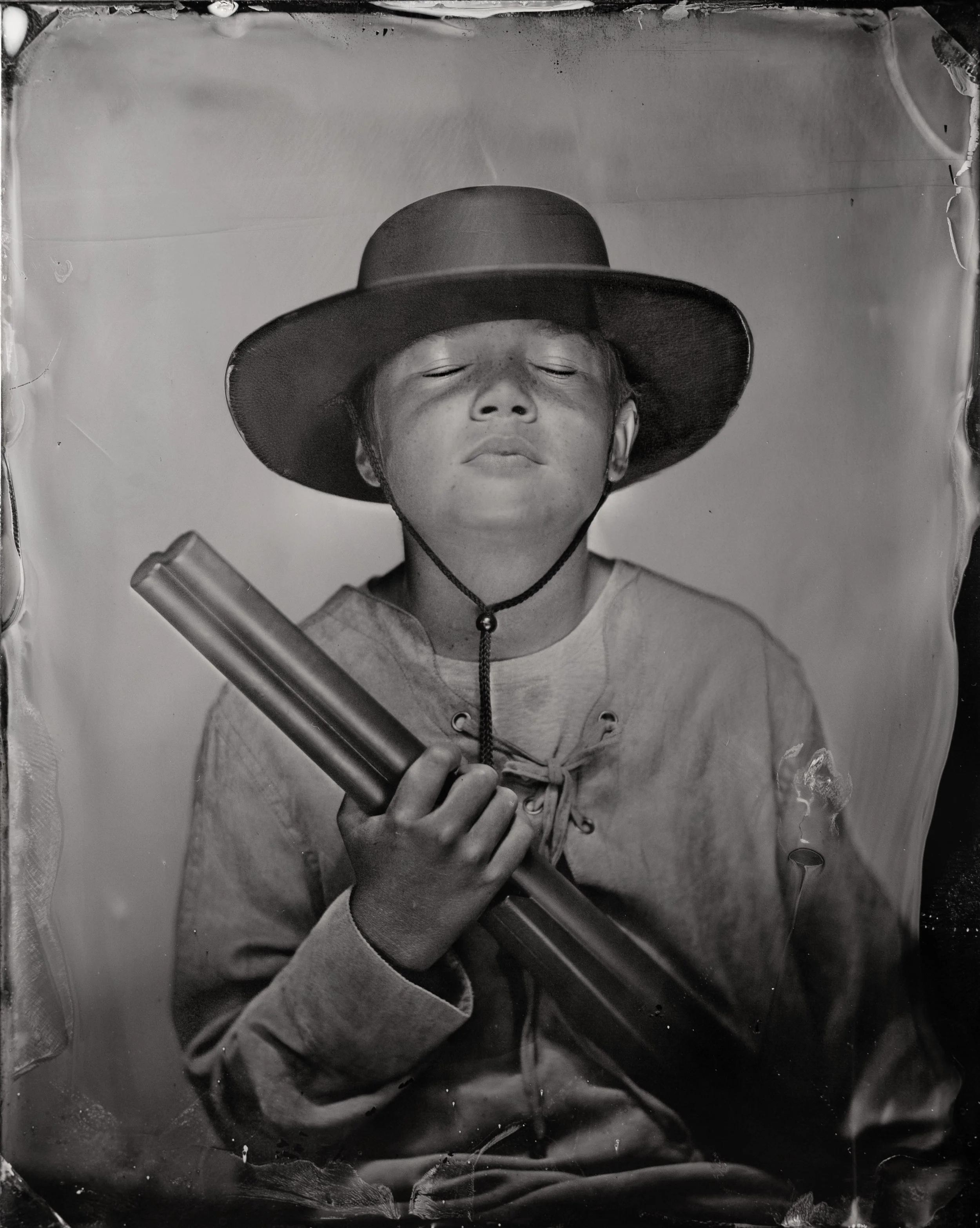 Black and white photograph of a boy wearing a wide-brimmed hat, holding a cylindrical object, with his eyes closed and a neutral expression.
