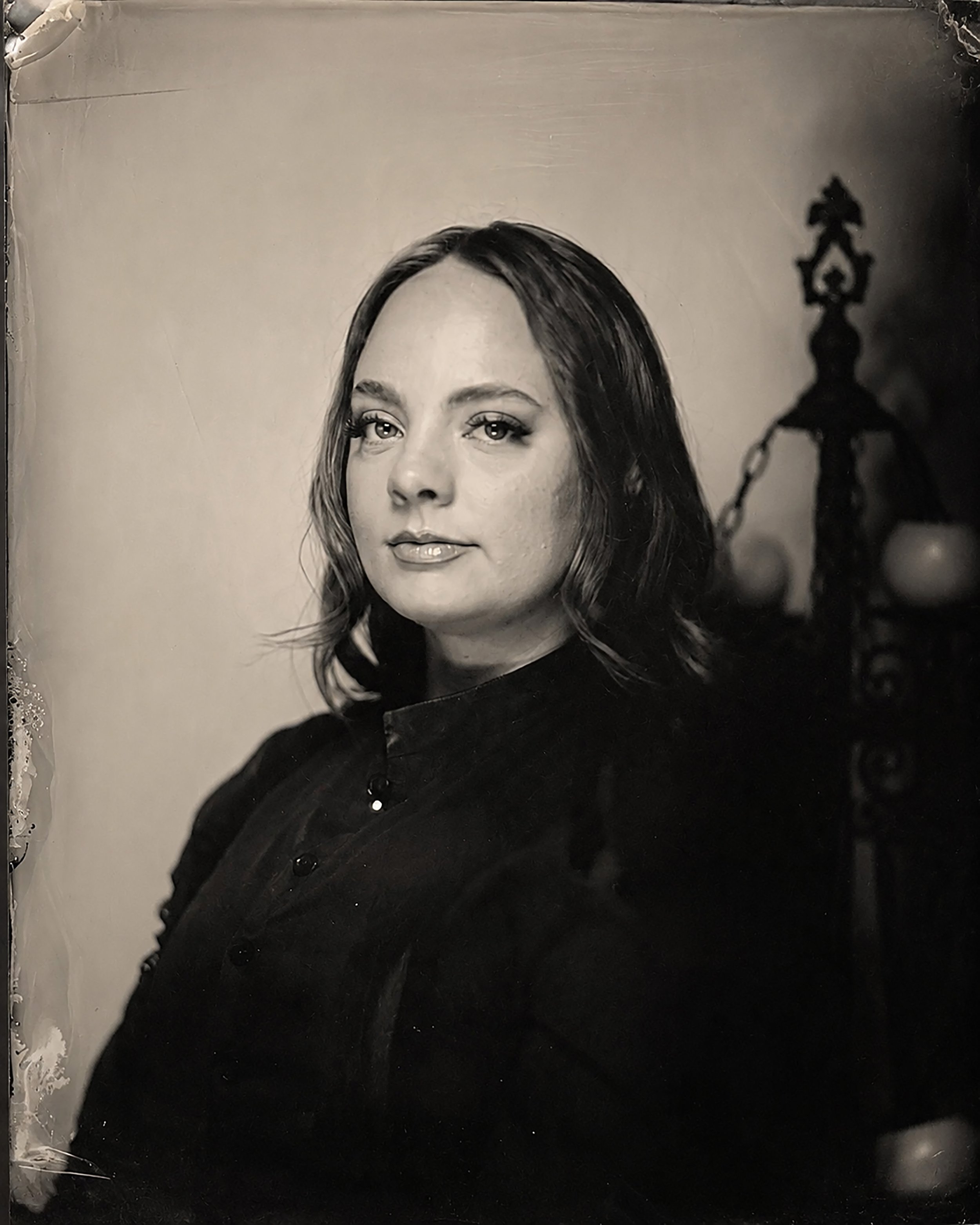 A black-and-white portrait of a woman with dark hair and light eyes, wearing a dark top, with a blurred background containing a decorative lamp.