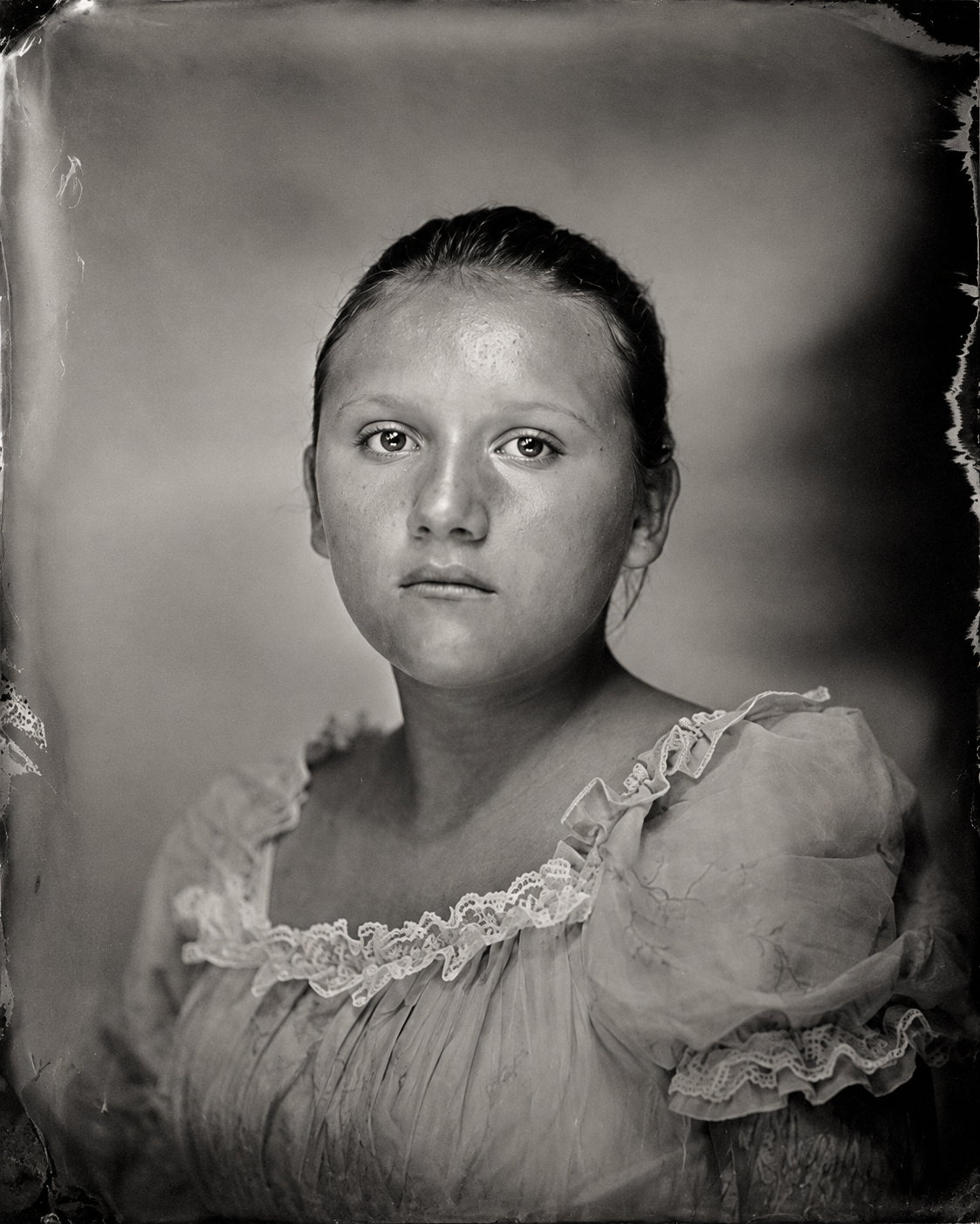 Black and white portrait of a young girl with slicked-back hair, wearing a dress with ruffled lace details around the neckline and sleeves.