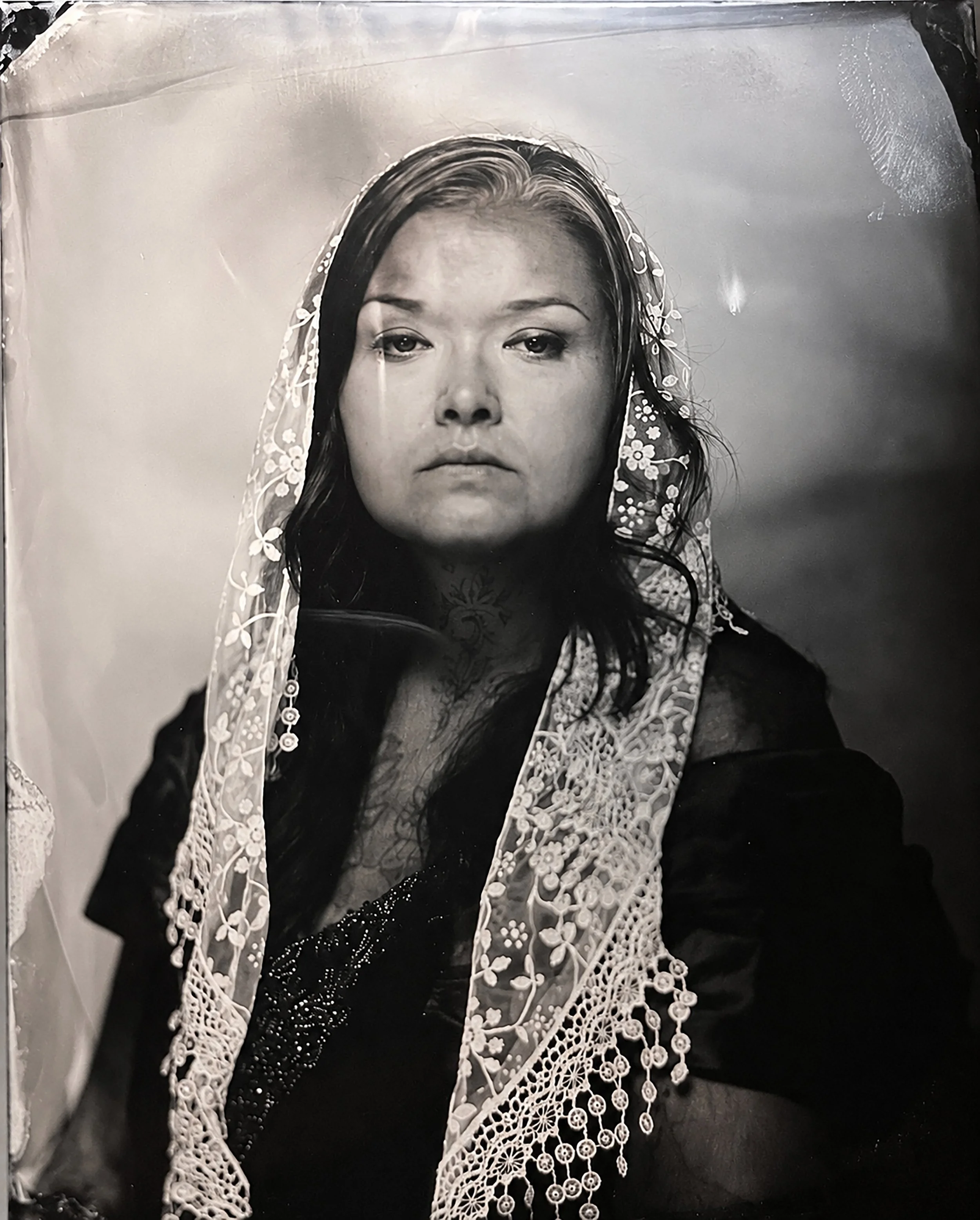 Black and white portrait of a woman with a serious expression, wearing a headscarf with floral embroidery and lace details, with long dark hair and a tattoo on her chest.  TINTYPE PORTRAIT