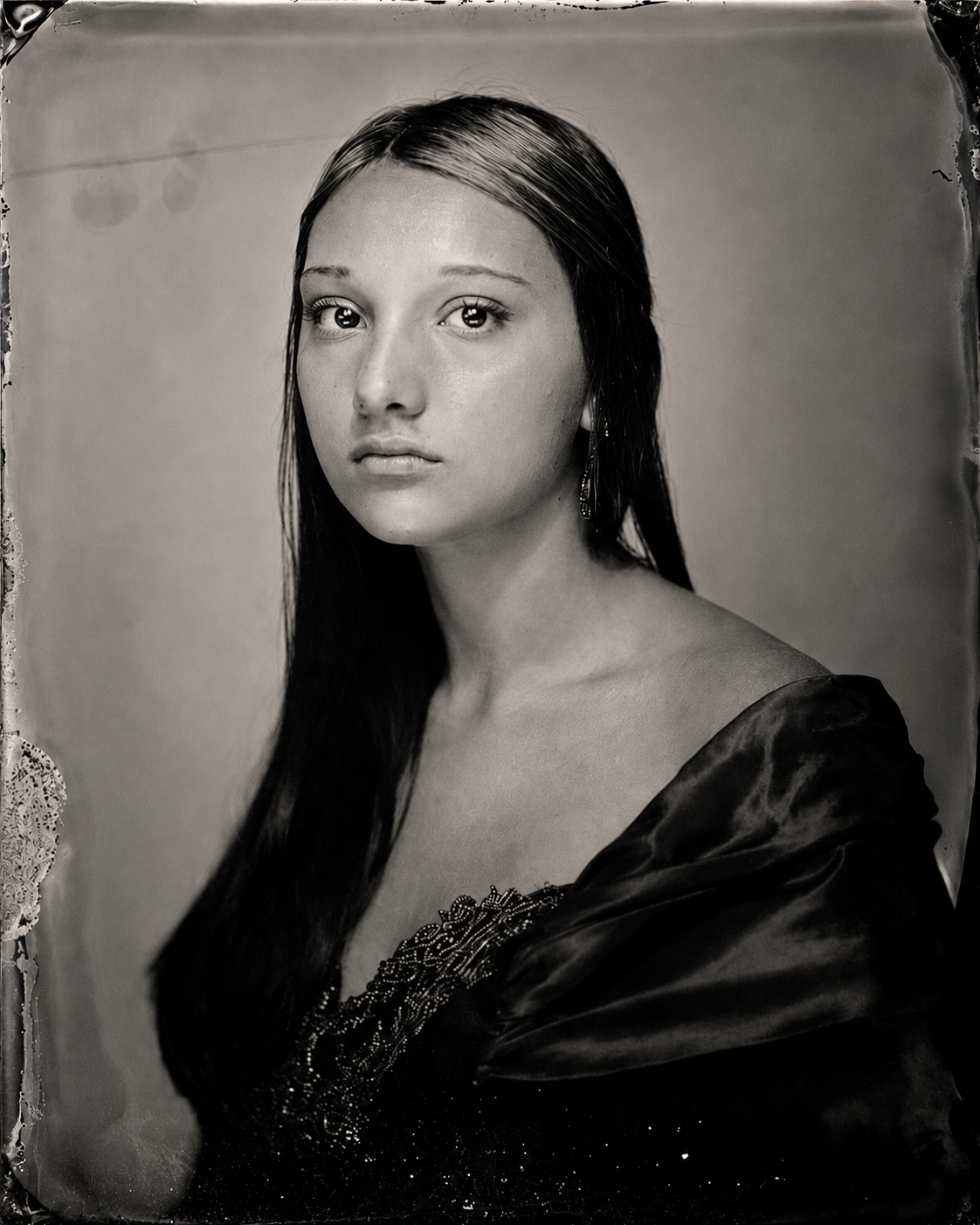 Black and white portrait of a young woman with long dark hair, wearing a dress with detailed embroidery and earrings, looking directly at the camera.
