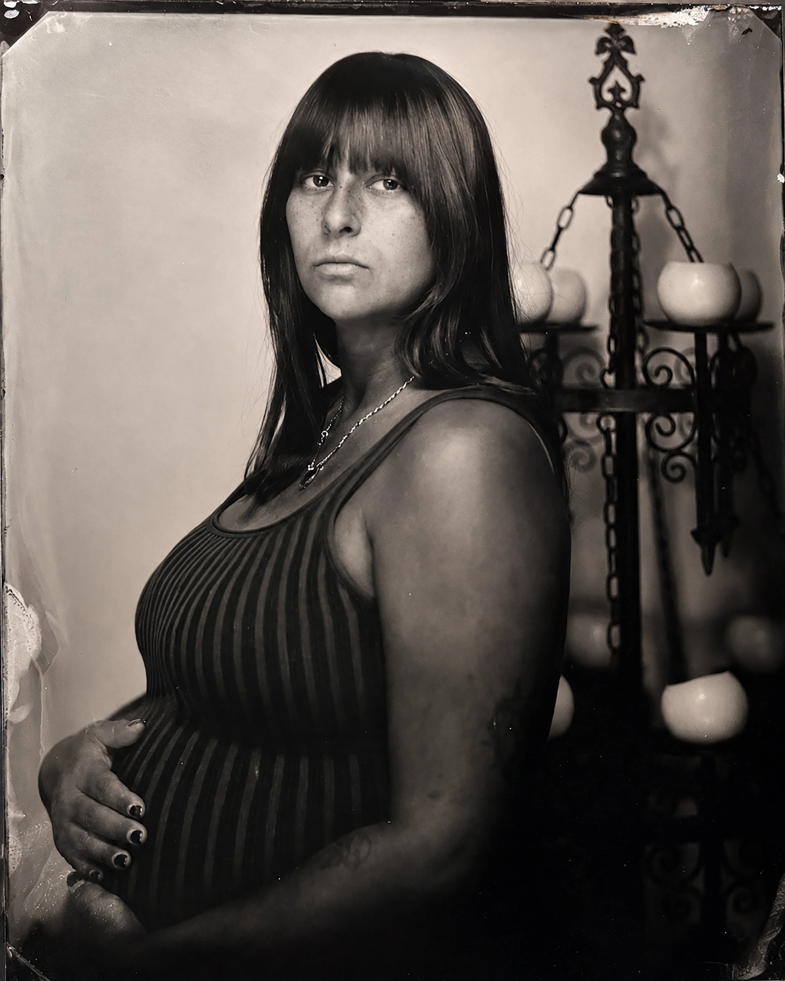 Black and white portrait of a woman with straight dark hair and freckles, wearing a striped top and necklace, standing next to an ornate candelabrum with unlit candles in the background.  TINTYPE PORTRAIT