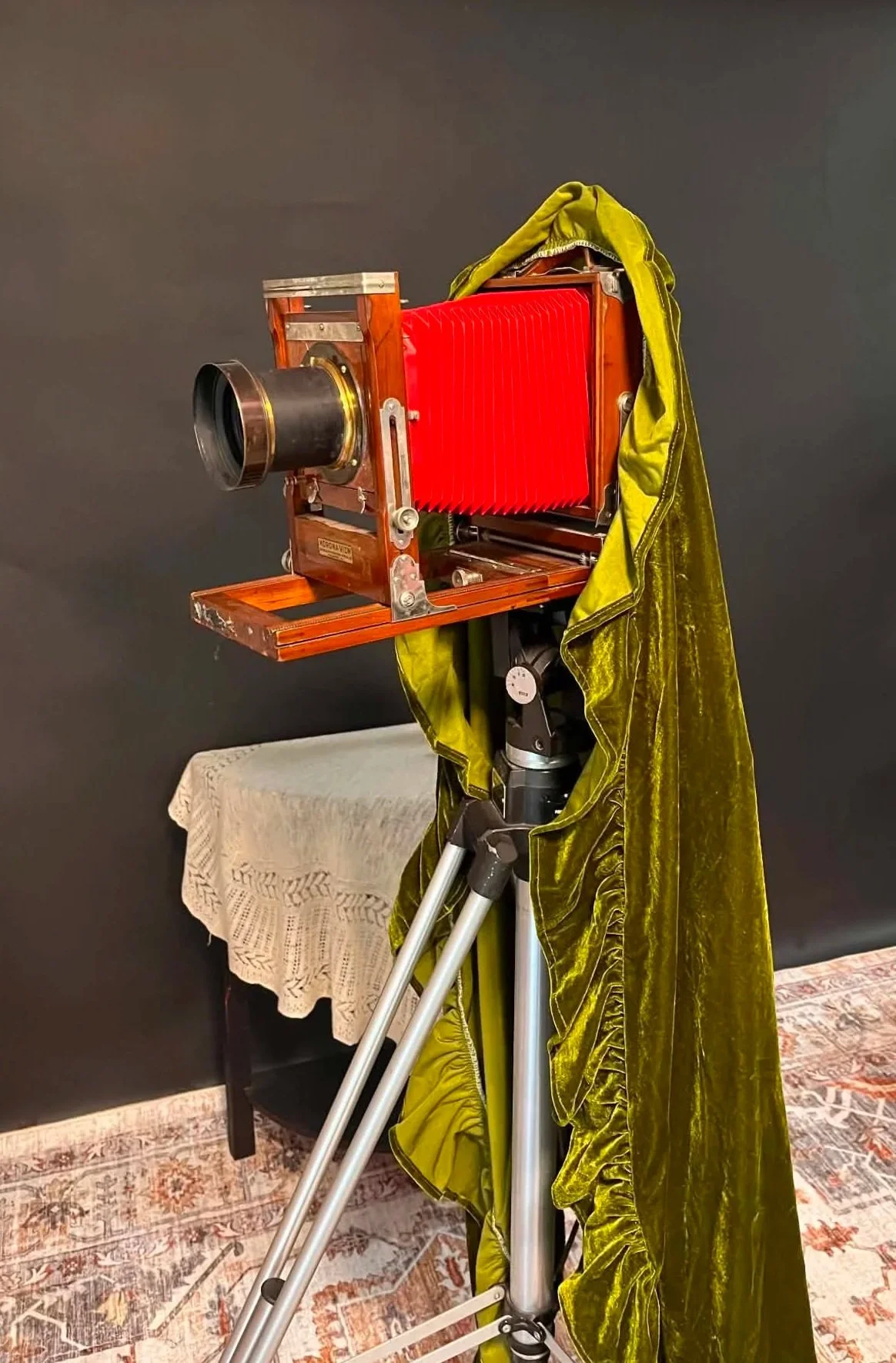 An old large-format camera mounted on a tripod, with a green velvet curtain draped over it, in a room with a dark wall, a table with a lace tablecloth, and a patterned rug.