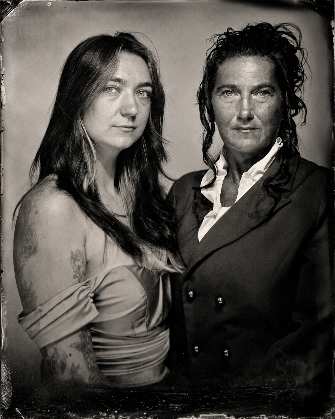 Black and white photograph of two women standing close together, both looking directly at the camera. The woman on the left has long dark hair, tattoos on her left arm, and is wearing an off-the-shoulder top. The woman on the right has curly hair, is