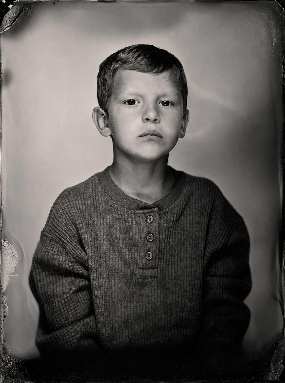 A black and white portrait of a young boy with short hair, wearing a buttoned, long-sleeve, knitted sweater.