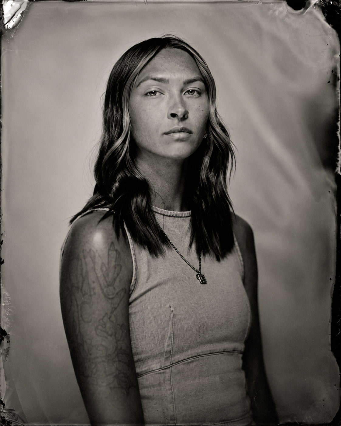 Black and white portrait of a woman with wavy hair, tattoos on her arm, wearing a sleeveless top and a necklace with a pendant, looking at the camera.