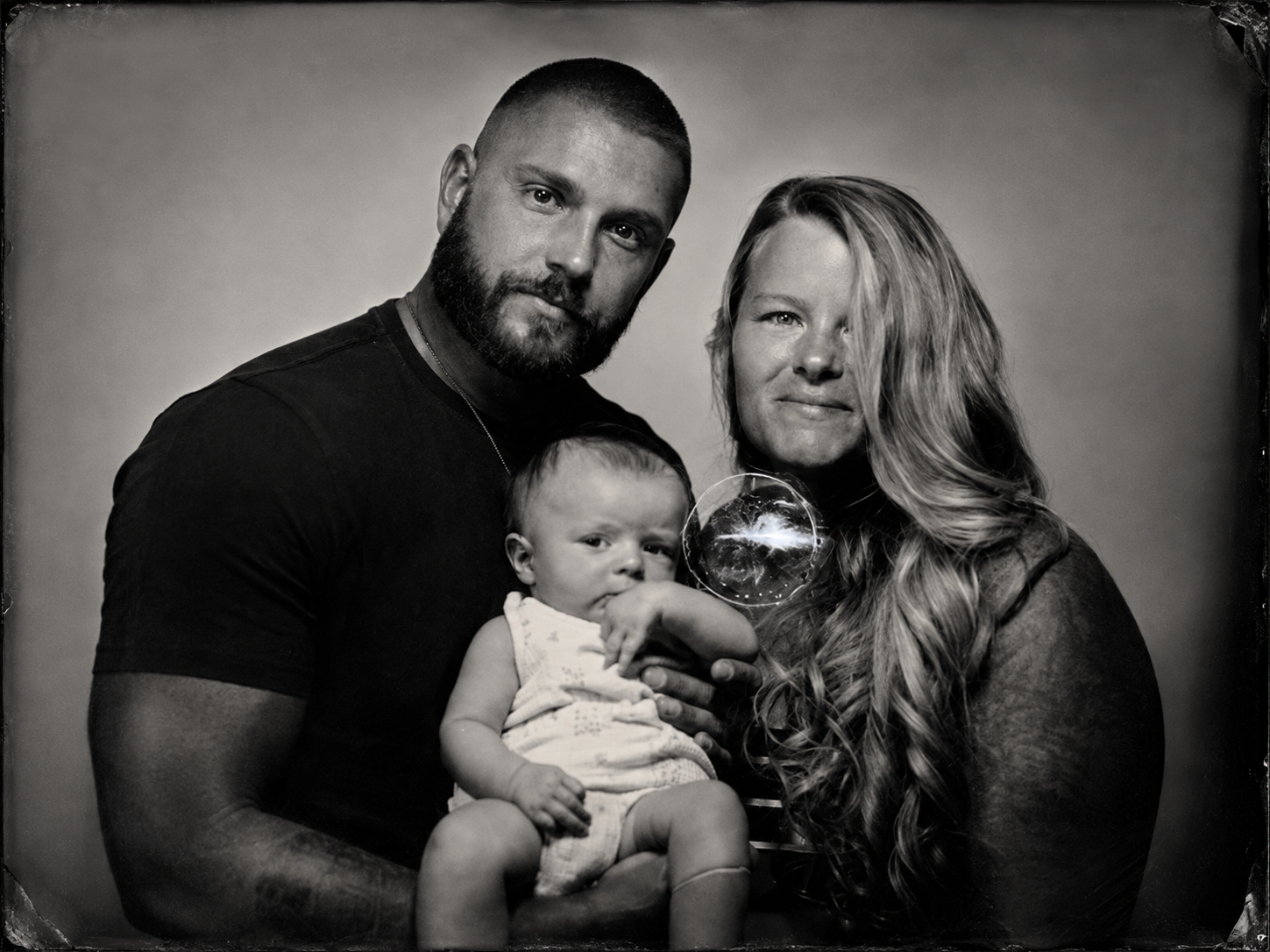 Black and white family portrait of a man, woman, and a baby, with the man holding the baby and the woman holding a bubble near her face.