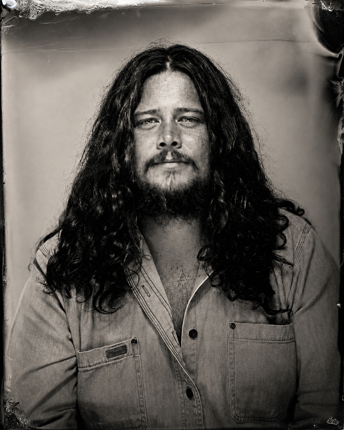 Black and white portrait of a man with long, curly hair and a beard, wearing a collared shirt, looking at the camera with a neutral expression.