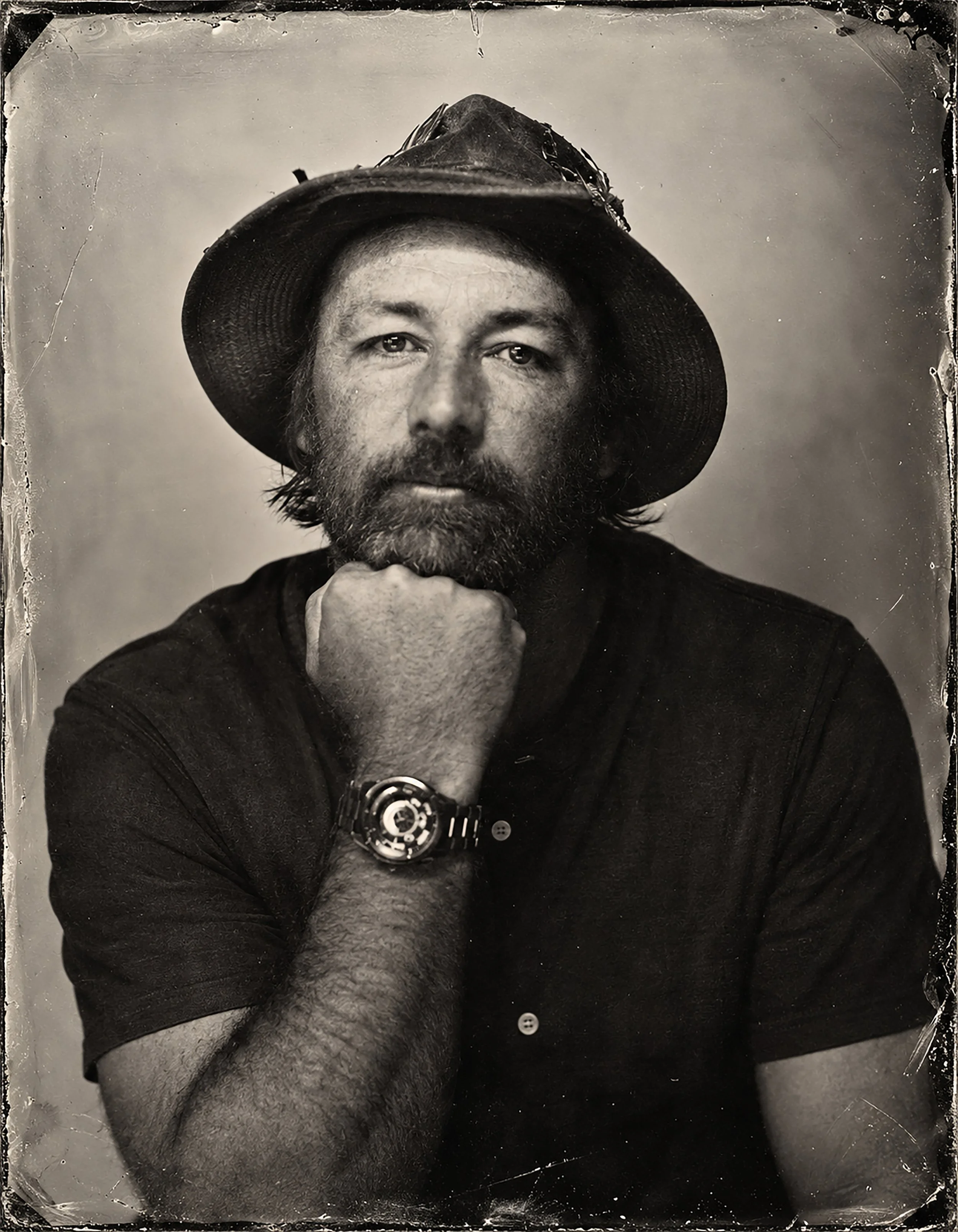 A man with a beard and mustache wearing a wide-brimmed hat decorated with various objects. He is resting his chin on his hand and wearing a wristwatch. The photo is in black and white with a vintage effect.