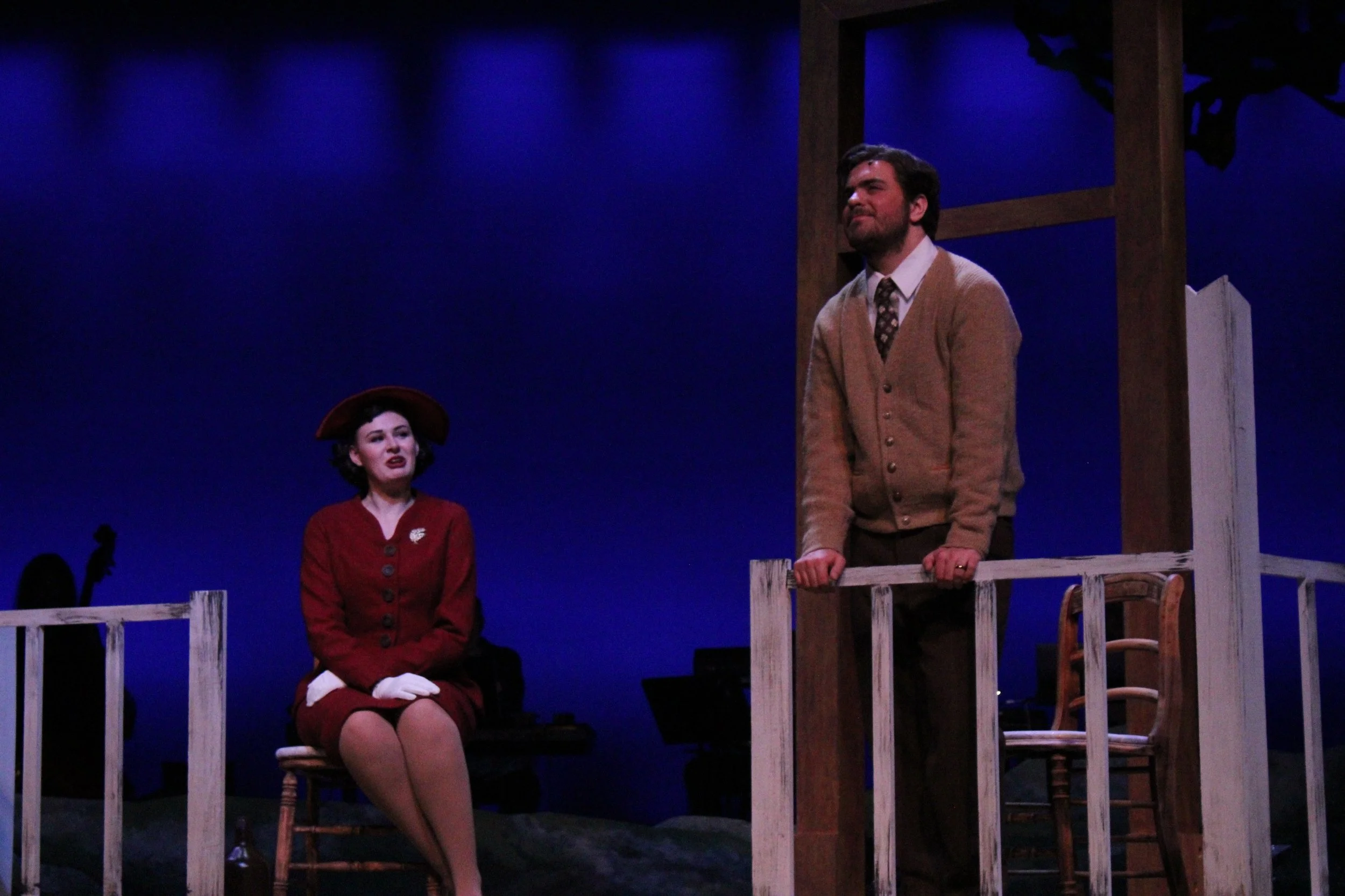 Two actors on stage, a woman sitting on a chair and a man standing at a railing, in front of a dark blue background with a wooden structure, during a theatrical performance.