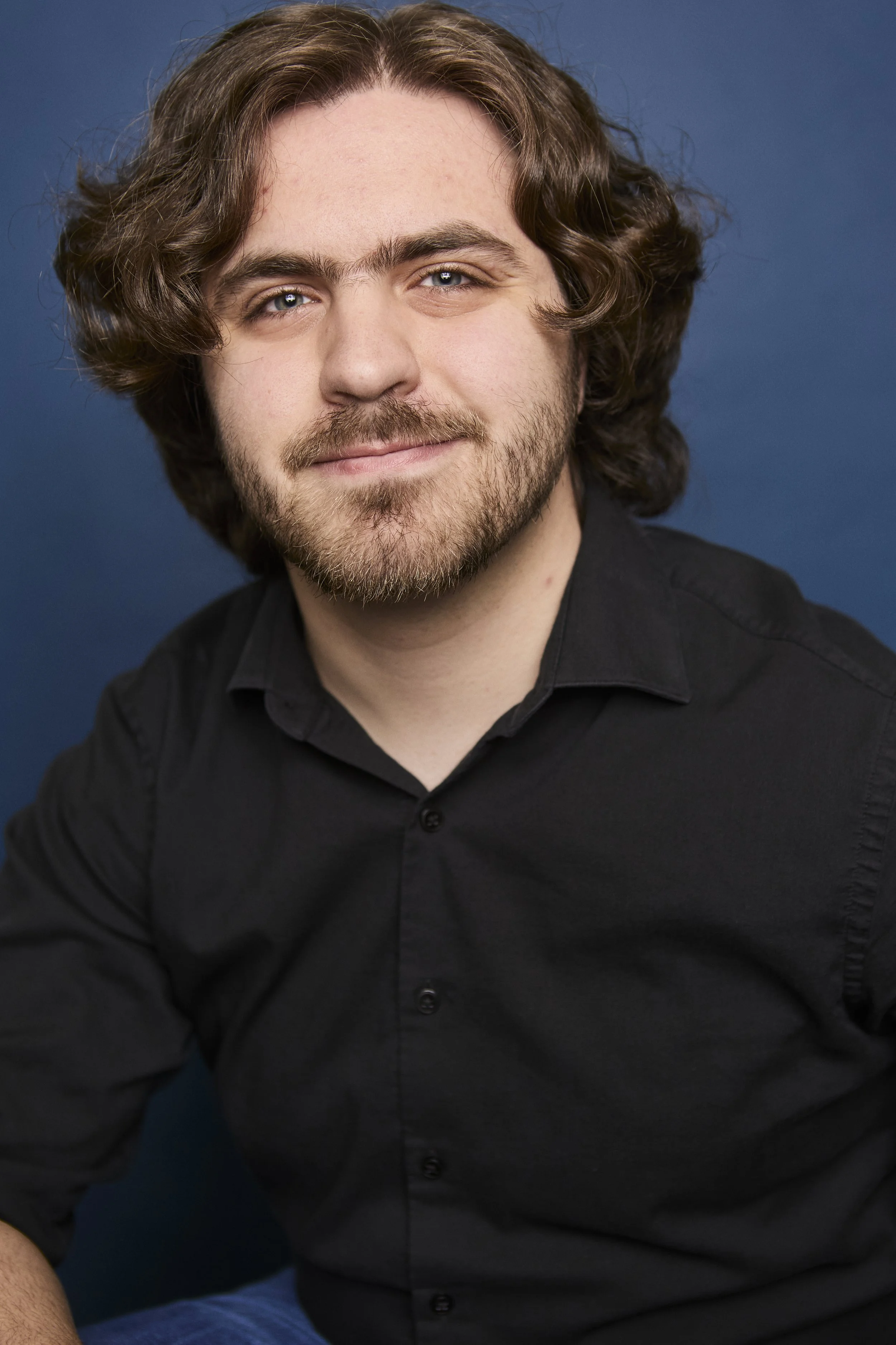 Close-up of a man with long curly brown hair and a beard, smiling while wearing a black shirt against a blue background.