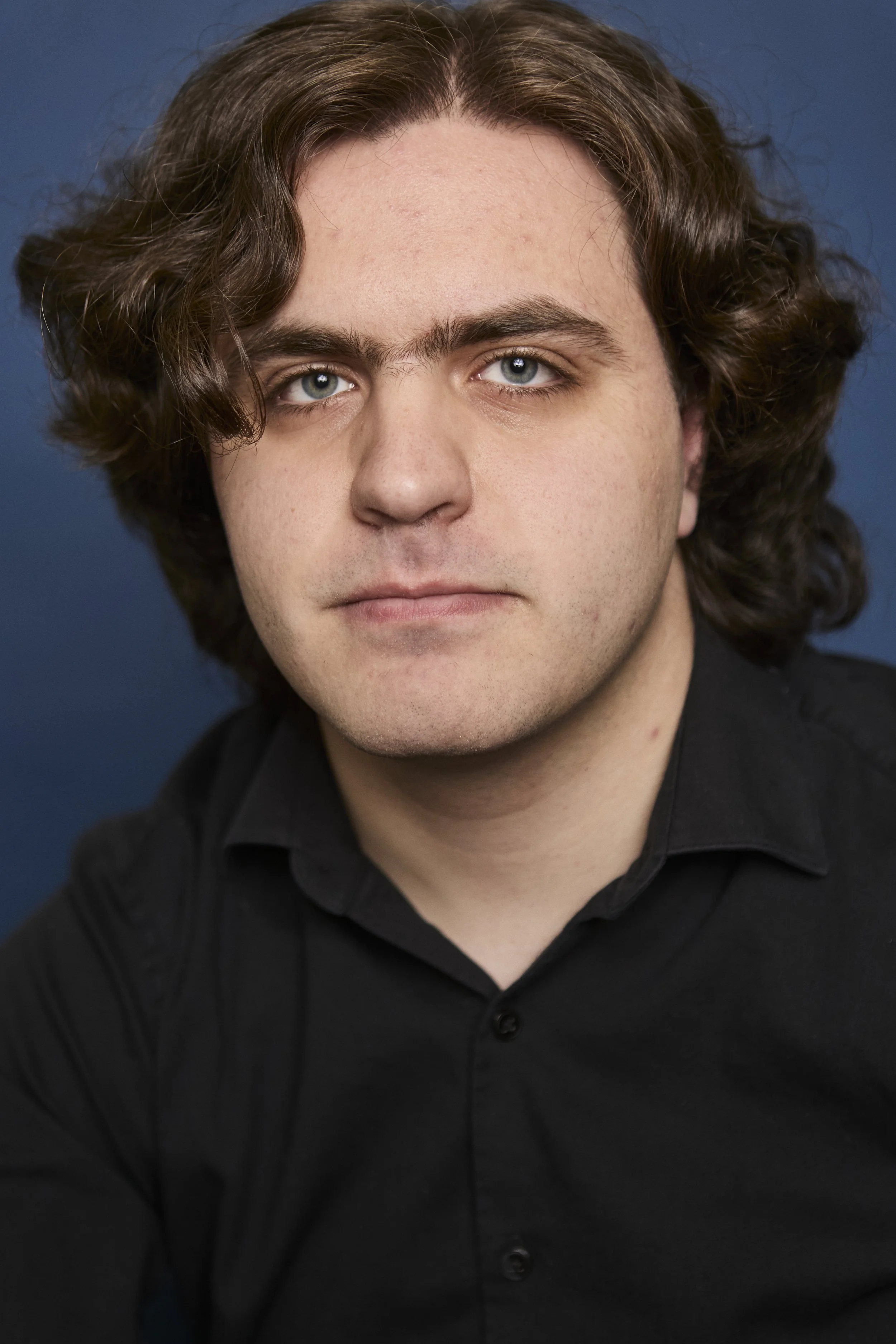 A close-up portrait of a young man with shoulder-length curly brown hair, light skin, and blue eyes, wearing a black collared shirt against a dark blue background.