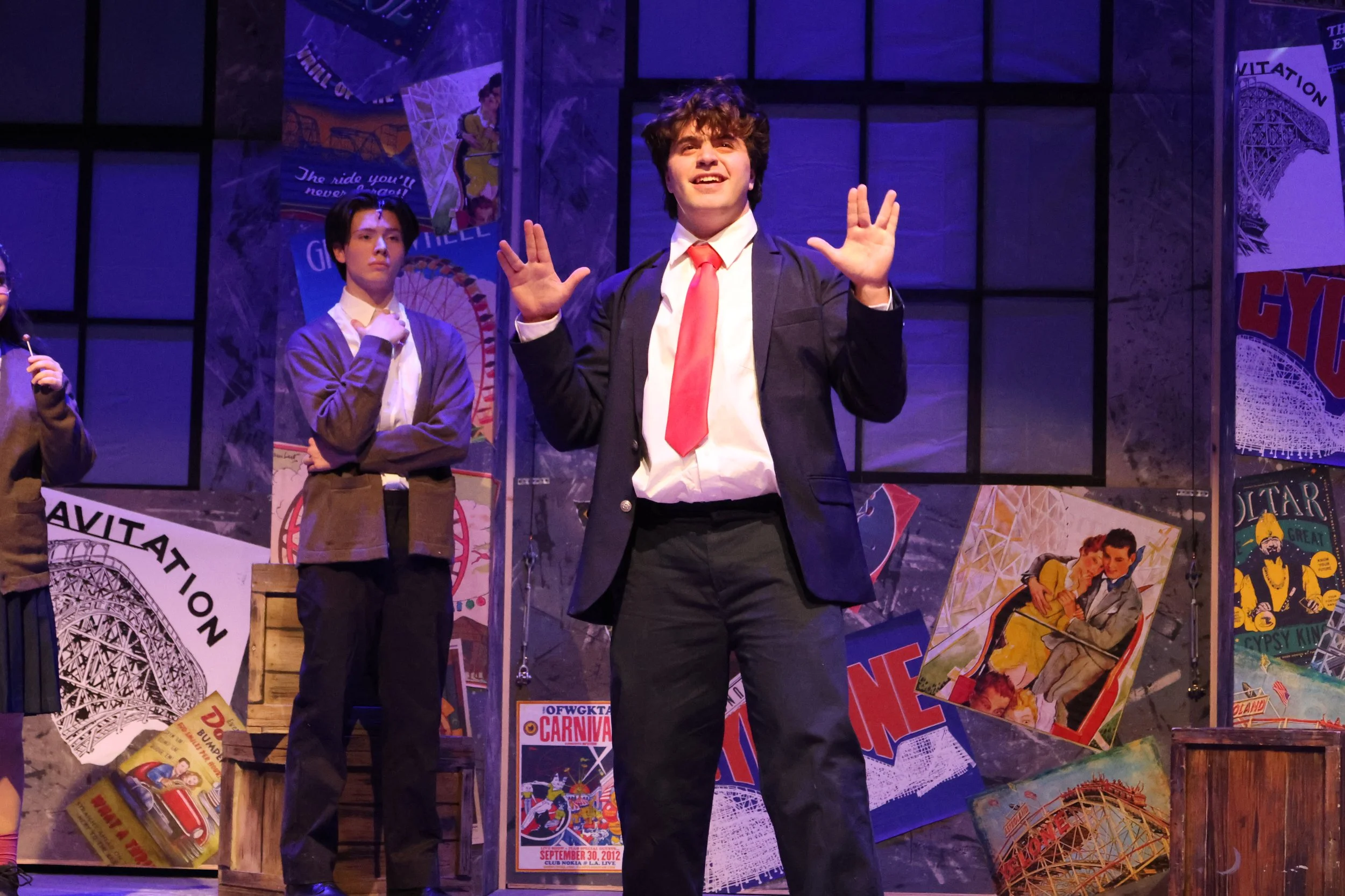 A group of young actors on a stage decorated with vintage carnival posters and images of amusement park rides. The central figure is a young man with curly hair, wearing a dark suit, white shirt, and red tie, smiling with his hands raised. The backgr