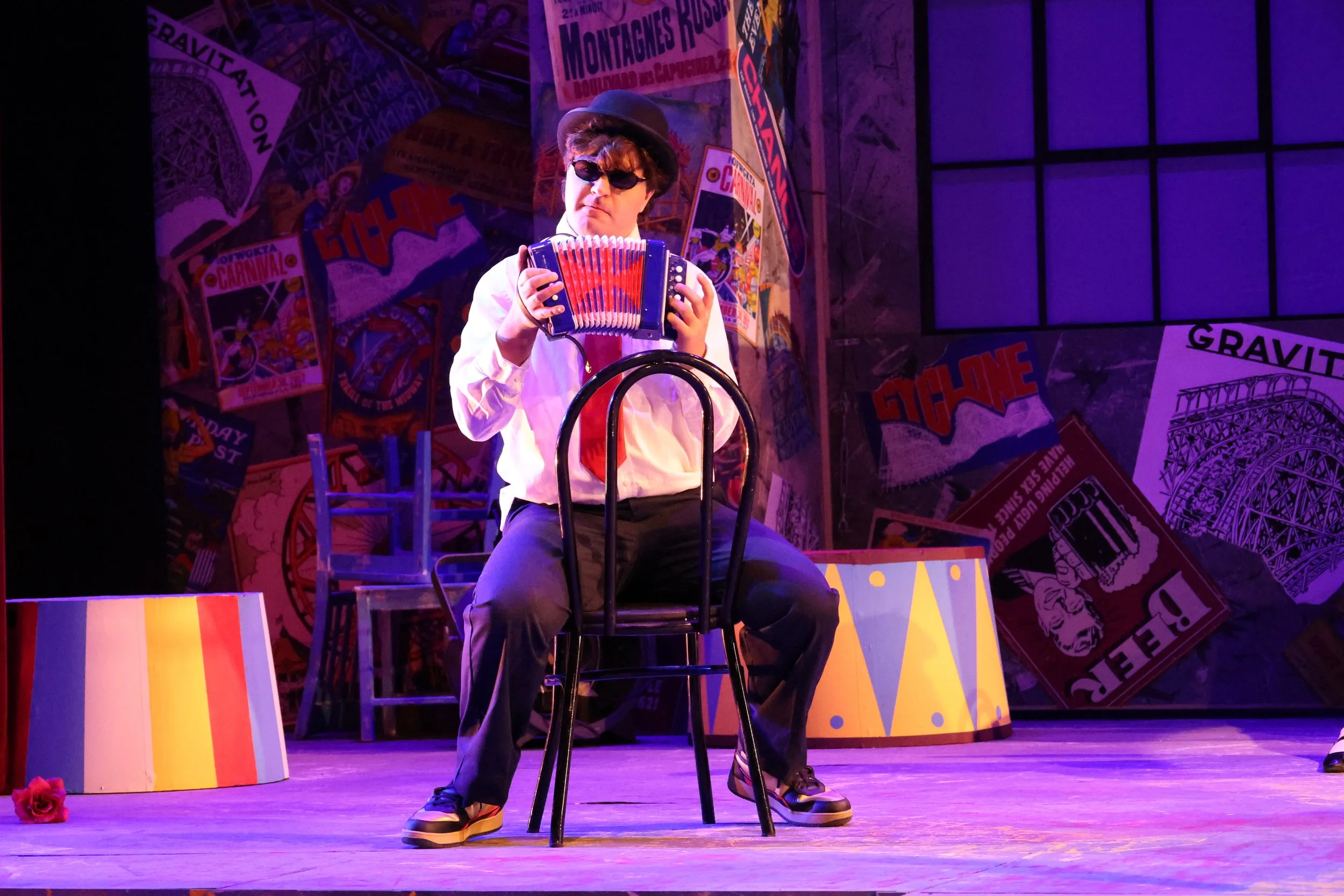 Performer sitting on a chair on stage, playing the accordion, wearing sunglasses, a hat, white shirt, and dark pants. The stage background features colorful posters and a purple lighting effect.