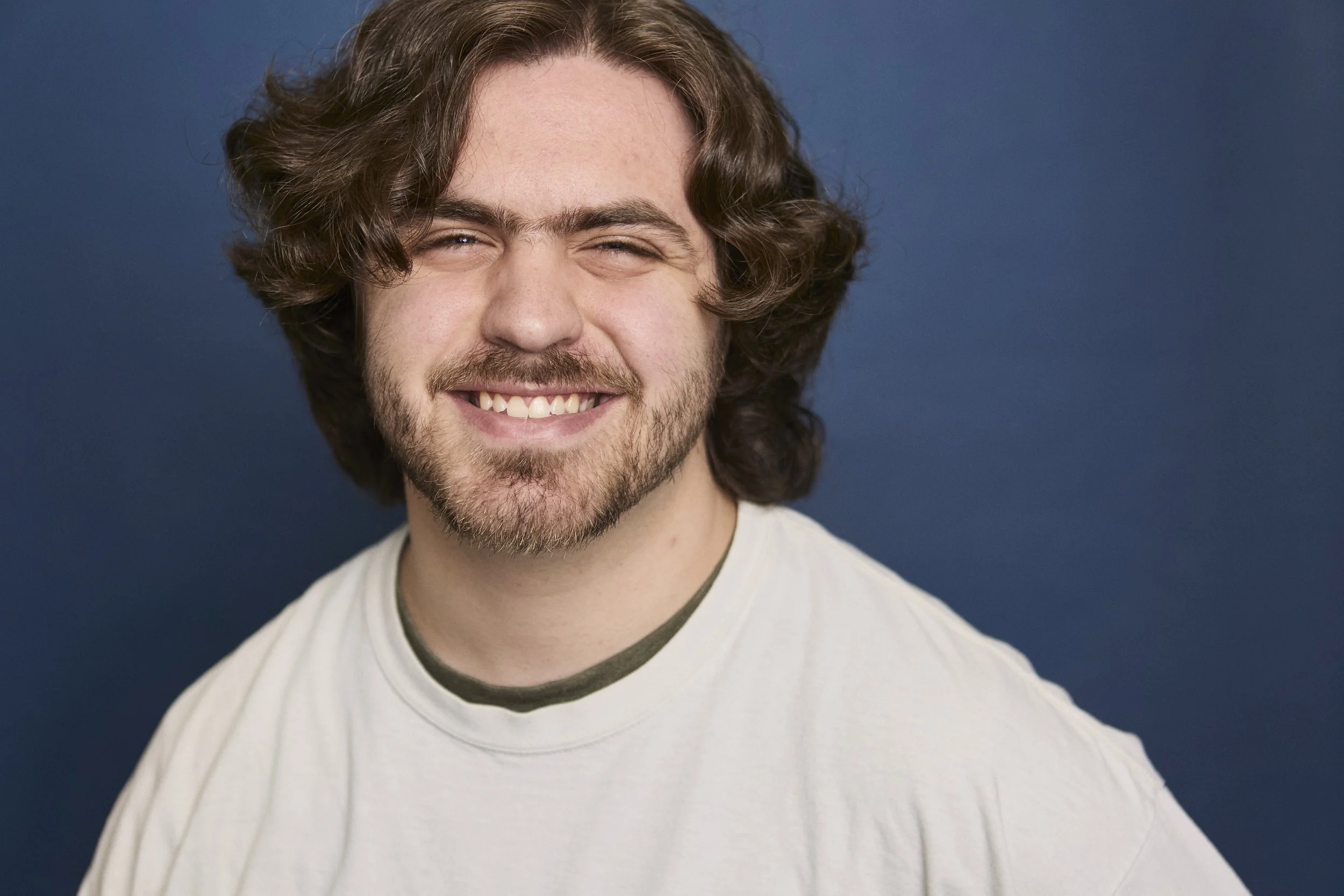 A young man with shoulder-length curly brown hair and a beard, smiling with a closed mouth, wearing a white t-shirt against a dark blue background.