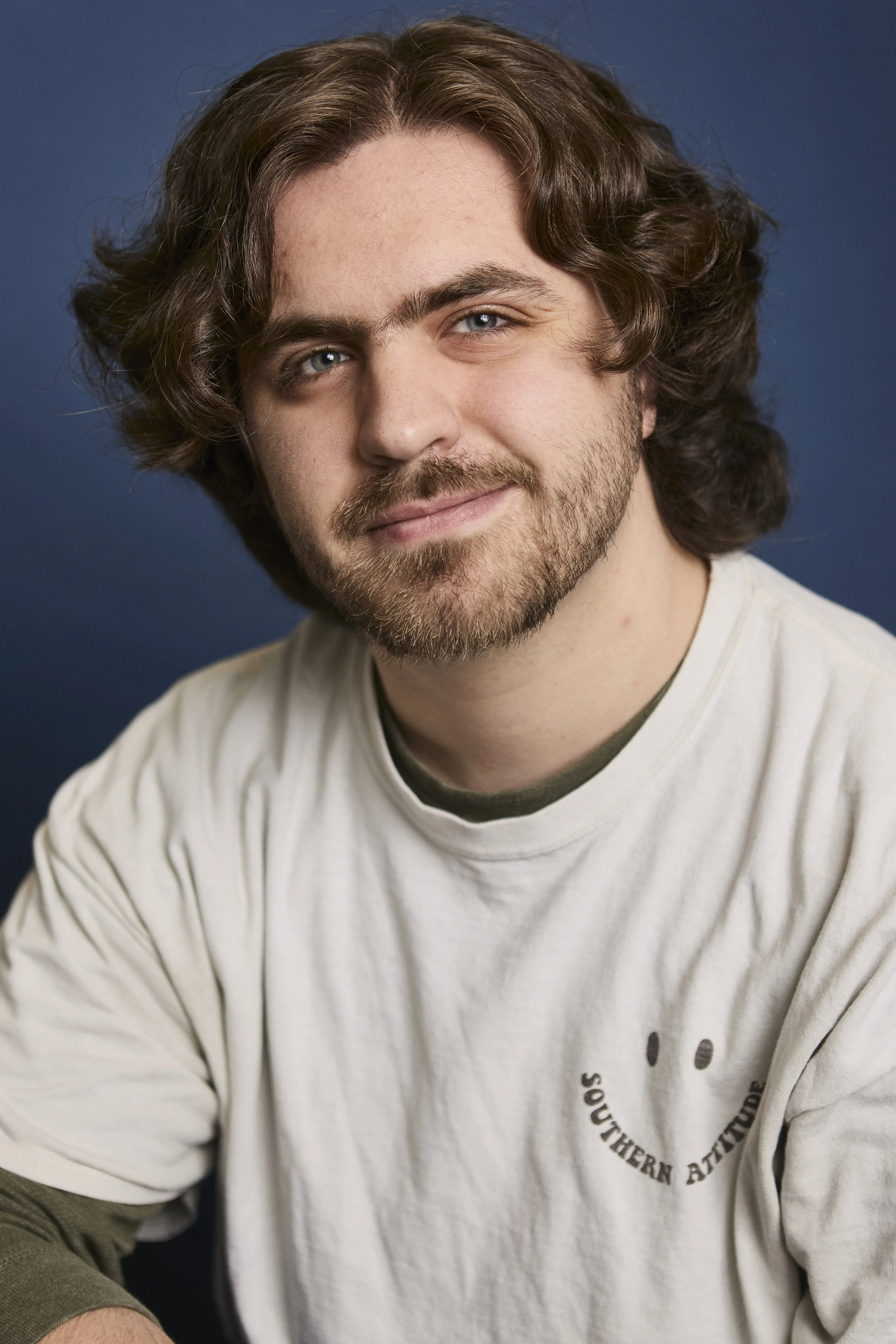 A man with medium-length wavy brown hair, light blue eyes, and facial hair wearing a light-colored t-shirt with a small dark-colored logo and text on it, against a dark blue background.