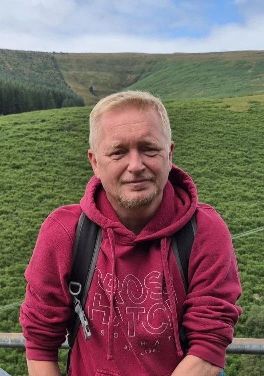A man with blond hair and a goatee, wearing a maroon hoodie and a backpack, standing outdoors on a hillside with lush green fields and rolling hills in the background.