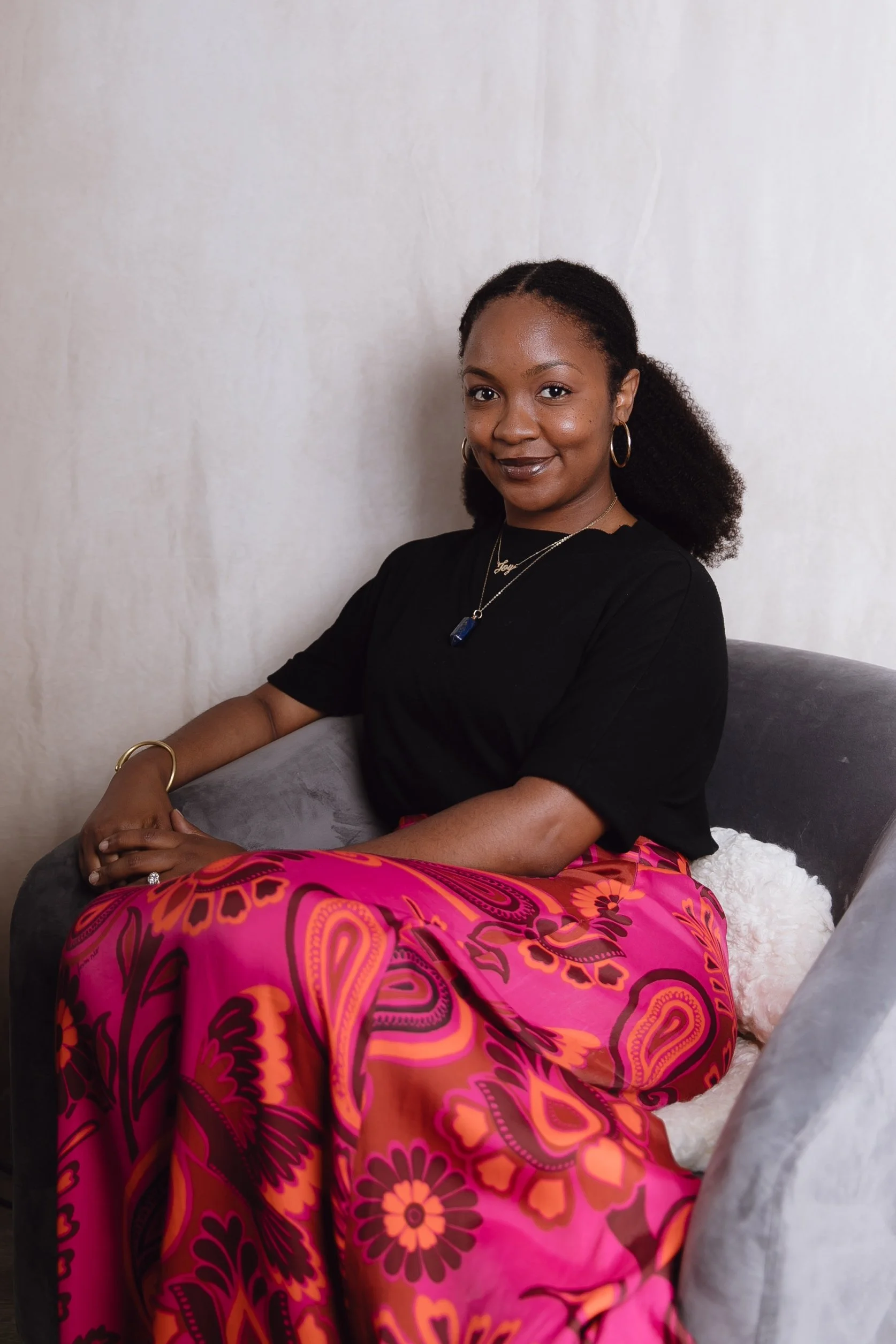 Angelica Joy Dumervil Perotti sitting on a gray velvet sofa, wearing a black top and vibrant pink patterned skirt, with her hands resting on her lap, against a plain light-colored wall.
