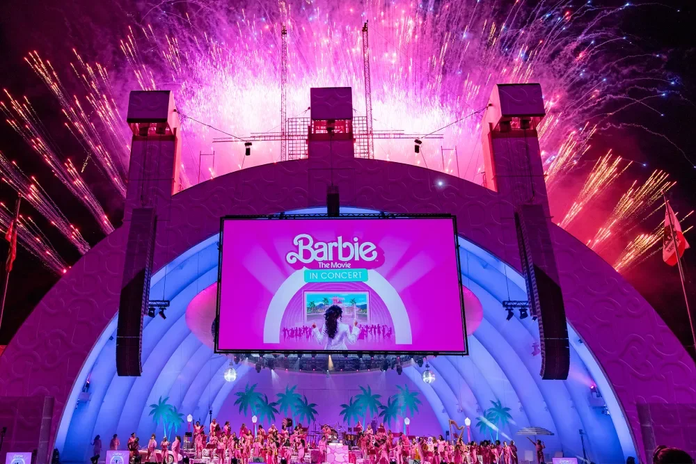 Colorful fireworks explode over an outdoor stage lit with pink and purple lights, hosting the 'Barbie' movie concert with a large screen displaying the Barbie movie's logo and a female performer in front of an orchestra at the Hollywood Bowl.