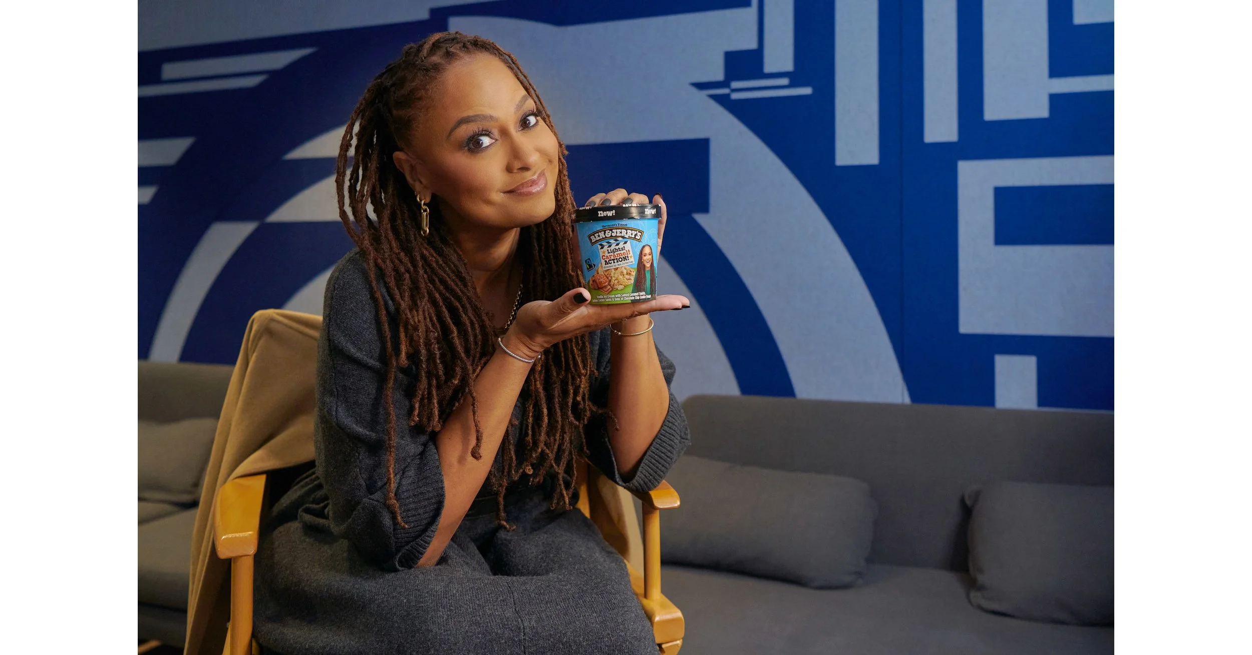 Ava Du Vernay with long, styled dreadlocks holding a tub of Ben & Jerry's ice cream and looking at the camera, sitting on a yellow chair in front of a blue wall with white geometric patterns.