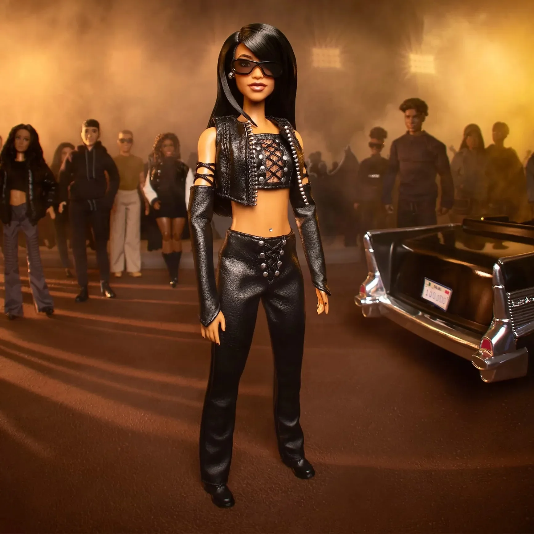 Aaliyah as a Barbie with long black hair wearing black leather pants, a black cropped top with lace-up detail, black gloves, and large sunglasses, standing in an indoor setting with a group of people in the background and warm lighting.