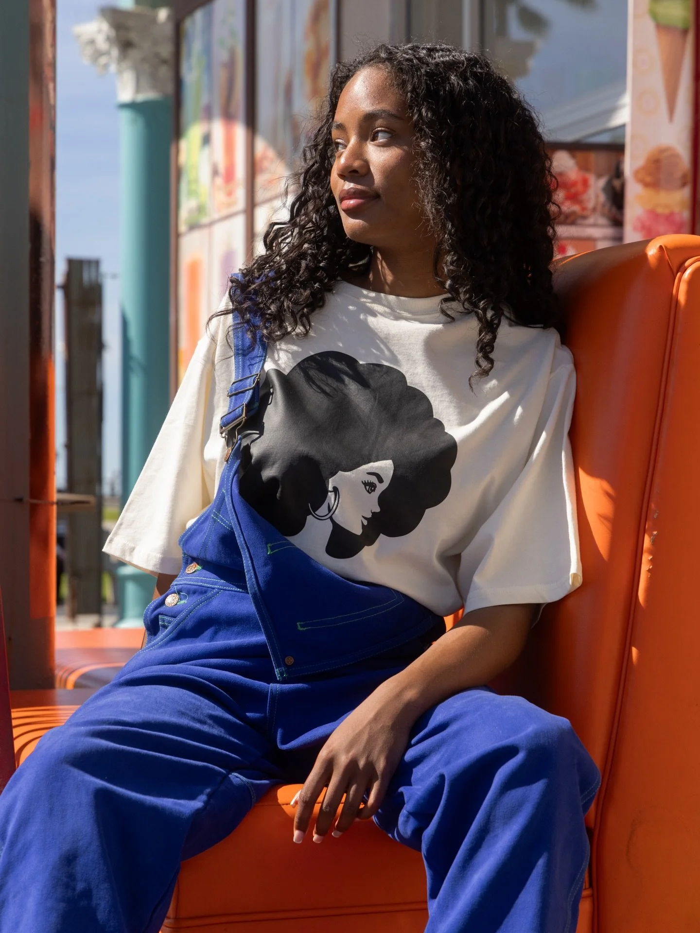 A young woman with curly hair wearing a white Cross Colours T-shirt with an illustration of a Barbie with an afro haircut, blue pants, and a blue bag, sitting on an orange bench outdoors on a sunny day.