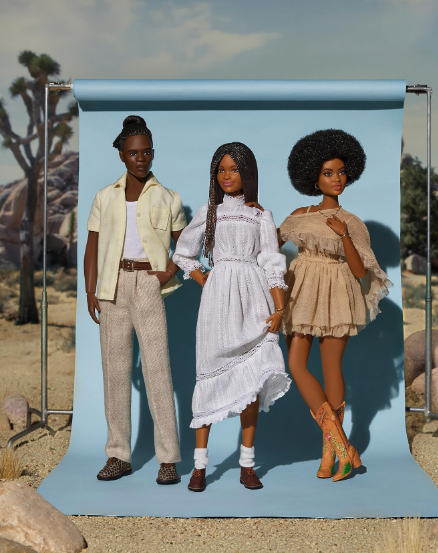Three Barbie dolls in fashionable outfits by Brother Vellies standing outdoors against a light blue backdrop with desert landscape, including rocks and Joshua trees.