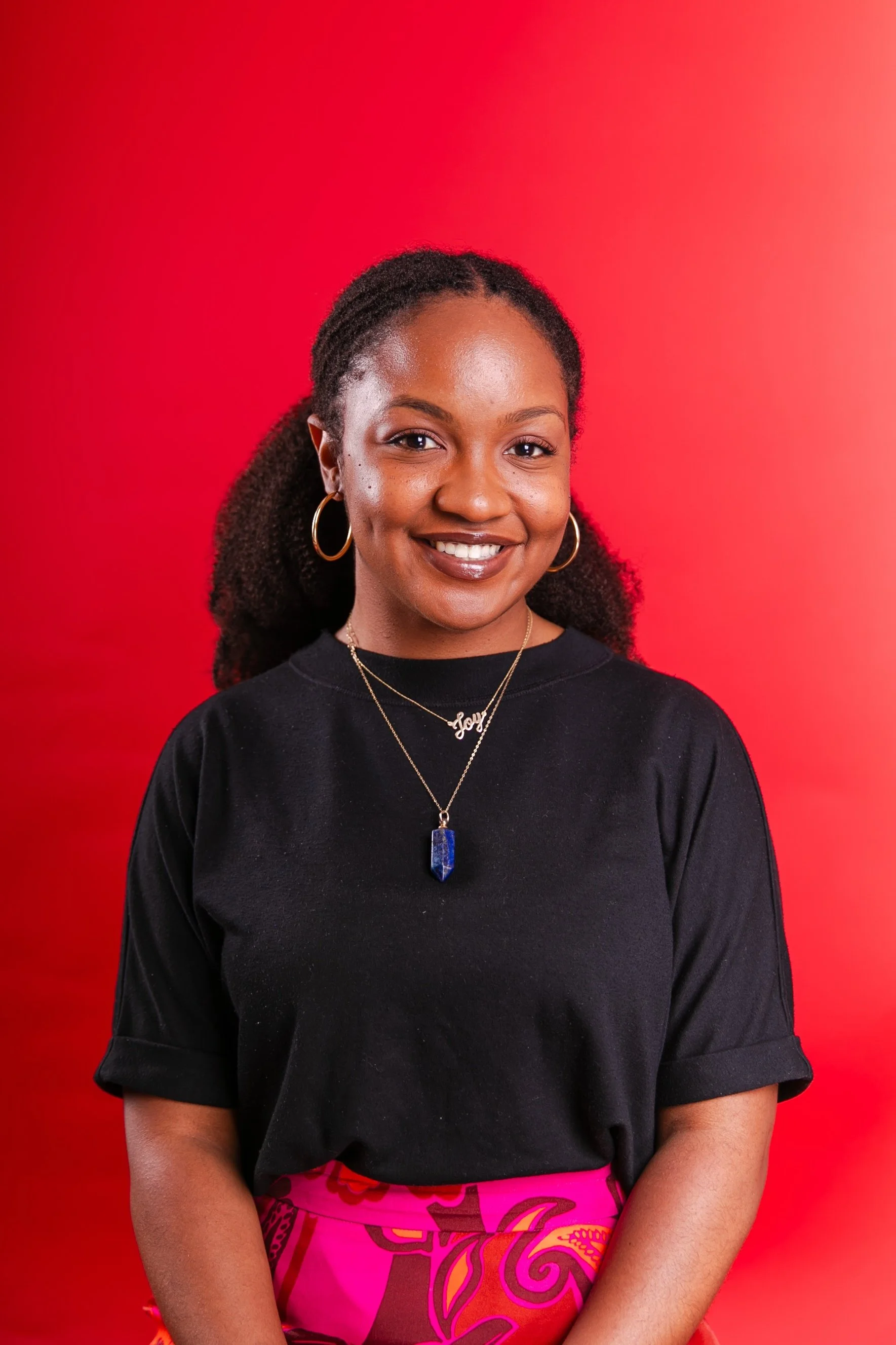 Angelica Joy Dumervil Perotti with dark curly hair, wearing a black top, gold hoop earrings, a gold necklace with the name 'Joy,' and a purple and pink patterned skirt, standing against a red background and smiling.