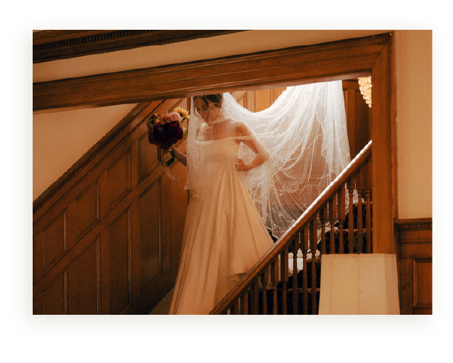 Bride holding a bouquet of flowers on a staircase, wearing a wedding dress and veil near a wooden railing and wall.