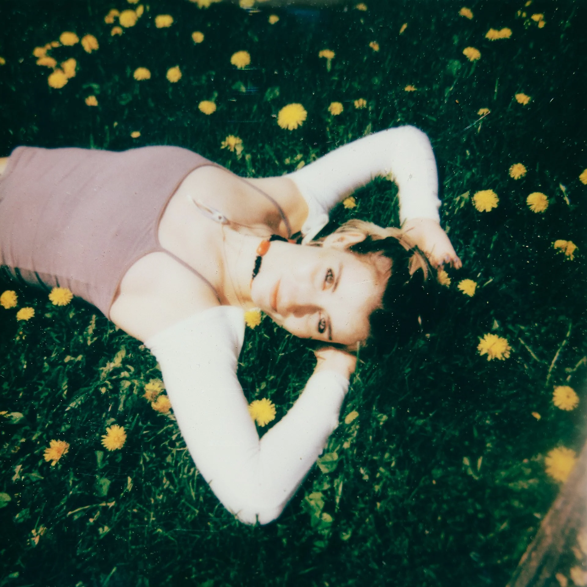 Young woman lying on grass near yellow dandelions, wearing a pink top and white pants, with blonde hair and glasses.