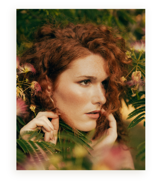 Close-up of a woman with curly red hair among green foliage and pink flowers, touching her face gently.