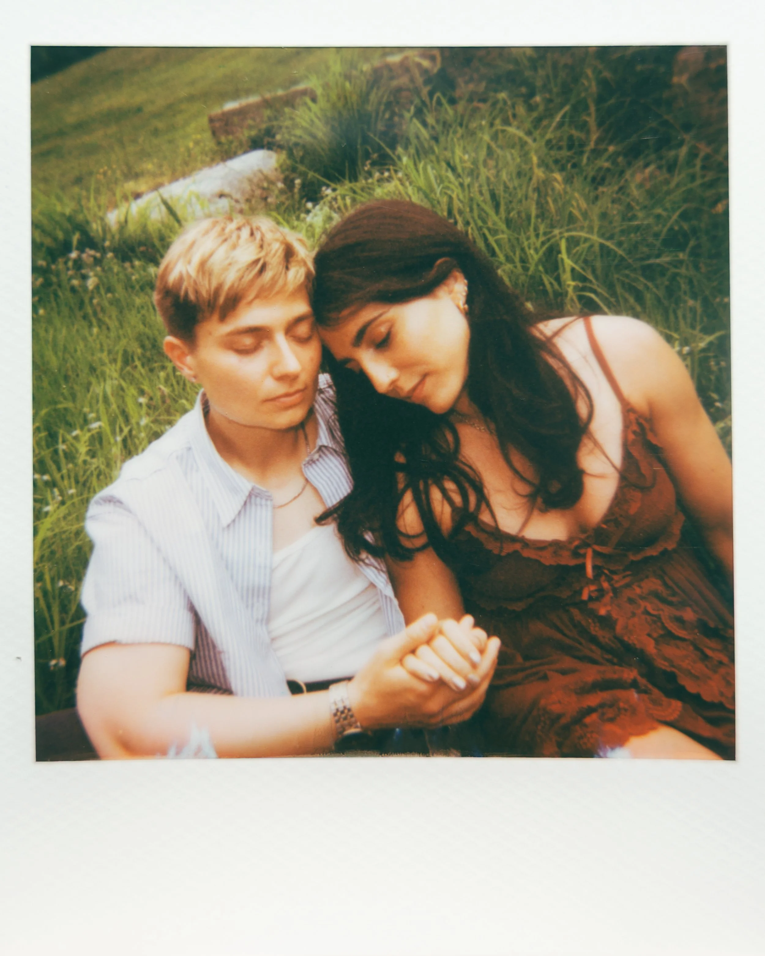 A young man and woman sitting closely together in tall green grass, with their eyes closed and heads touching, holding hands.